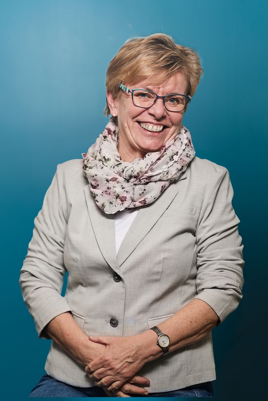 Ältere Frau mit kurzer blonder Frisur, Brille mit buntem Gestell, einem hellgrauen Sakko, floral gemustertem Tuch und silberner Armbanduhr lächelt freundlich vor blauem Hintergrund.