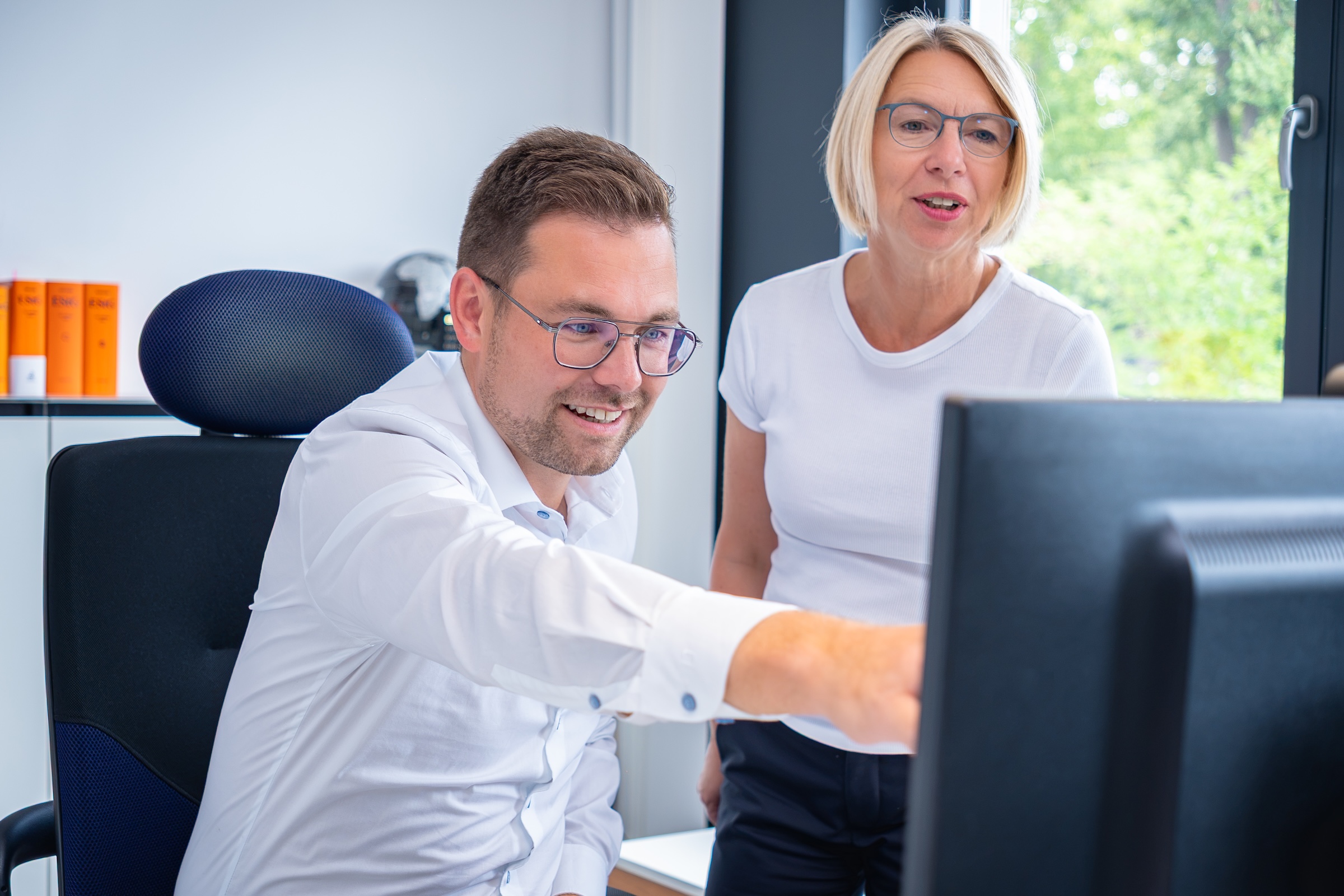 Ein Mann mit Brille und ein weißhaarige Frau schauen gemeinsam auf einen Computermonitor in einem hellen Büro mit Blick auf Grünfläche draußen.