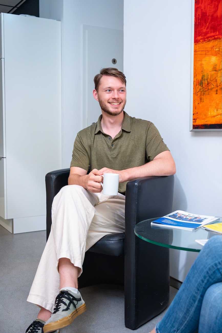 Junger Mann in grünem Poloshirt und beiger Hose sitzt lächelnd in einem schwarzen Sessel, hält eine weiße Tasse, an einem Glastisch mit Zeitschriften an der Seite.