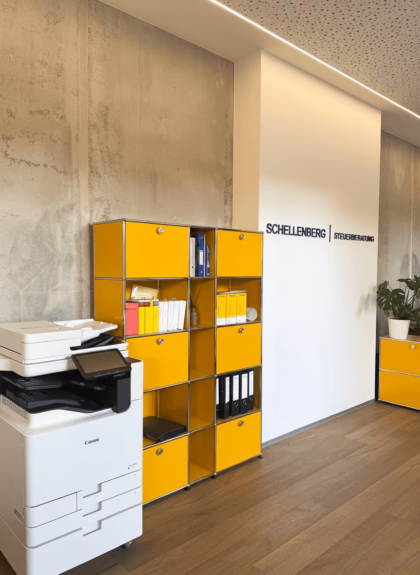 Moderner Bürobereich der Schellenberg Steuerberatung mit gelben Aufbewahrungsmodulen, Drucker und Kanzleischriftzug an der weißen Wand.