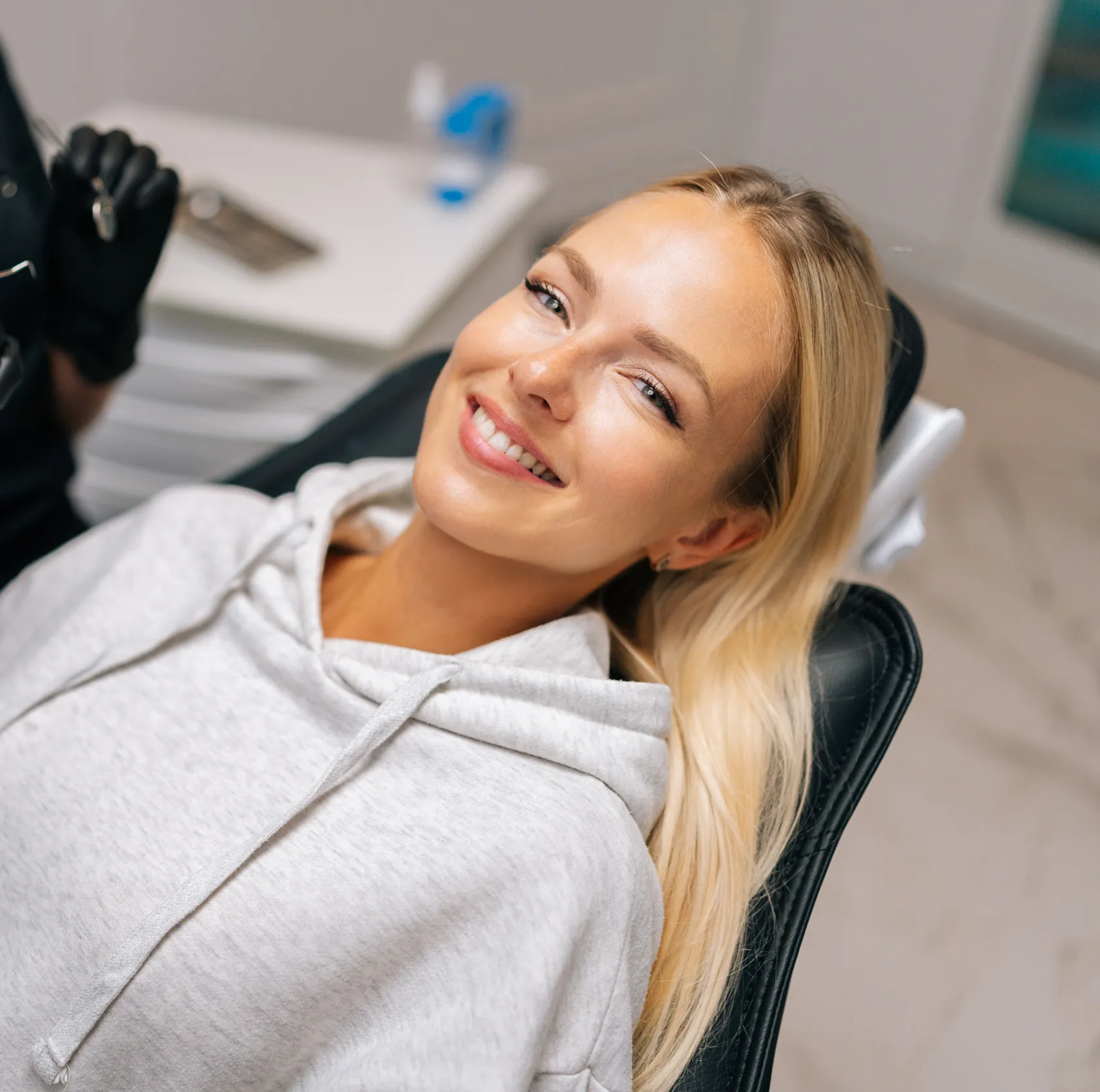 Smiling woman with blonde hair reclining in a dental chair, wearing a light gray hoodie.