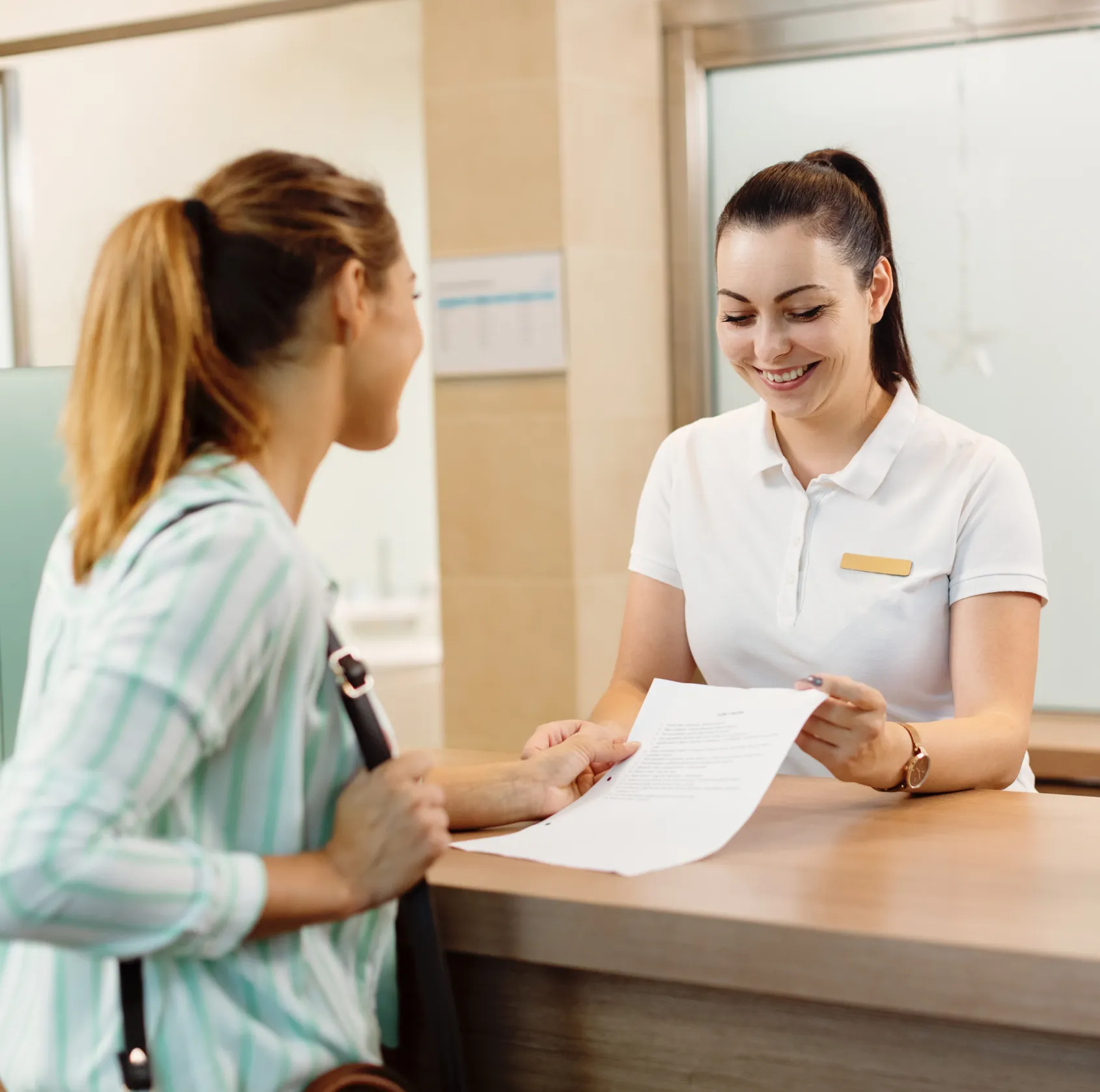 Receptionist smiling while handing a document to a woman at the front desk.