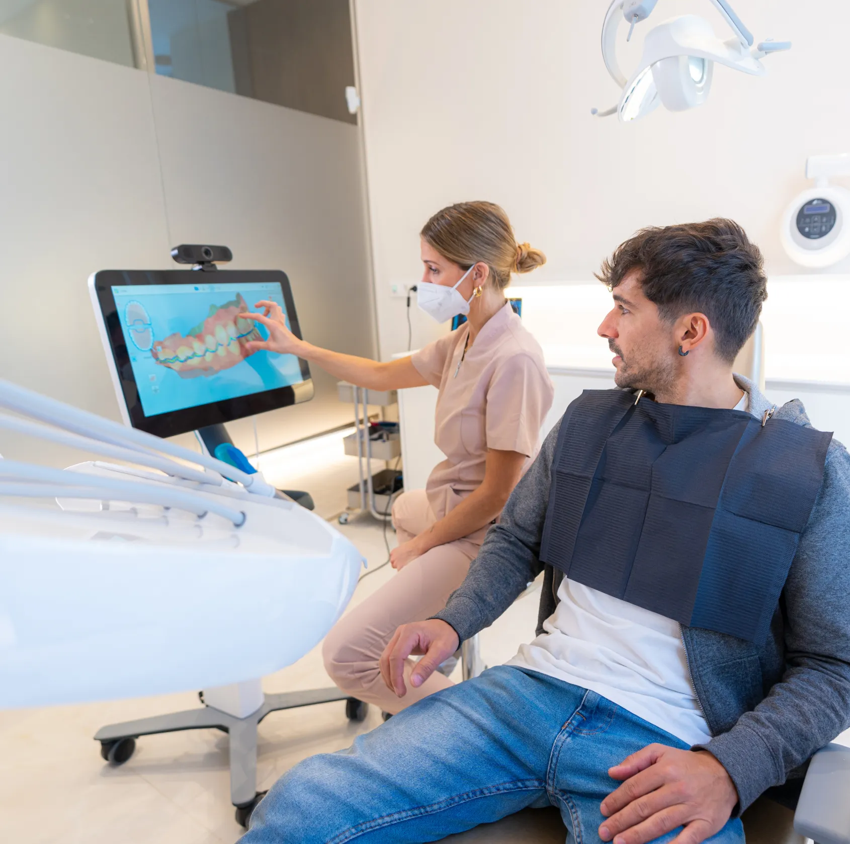 Dentist in mask showing dental scan on screen to a seated male patient with dental bib.