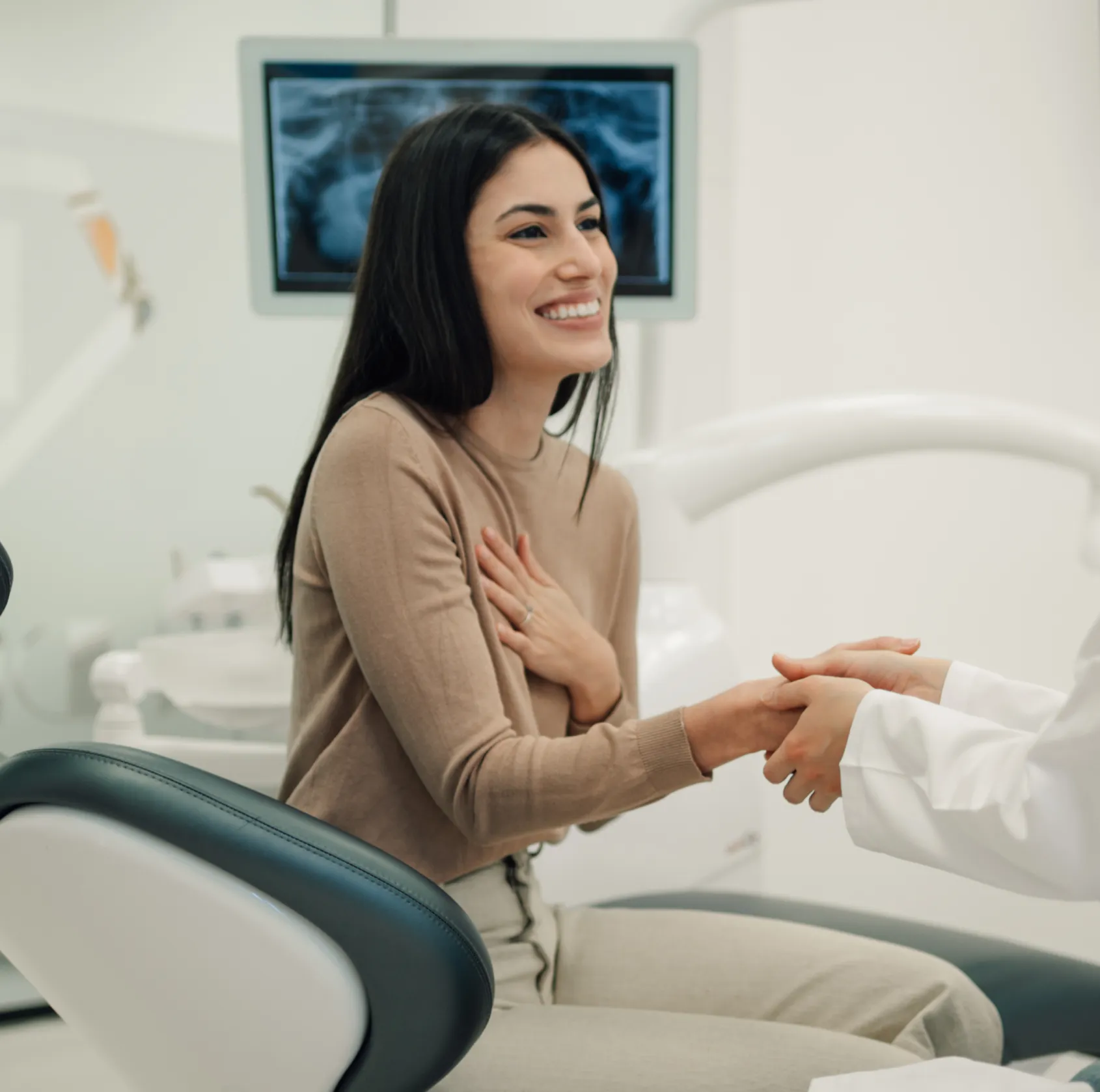 Smiling woman sitting in dental chair holding dentist's hand with dental X-ray on screen behind.