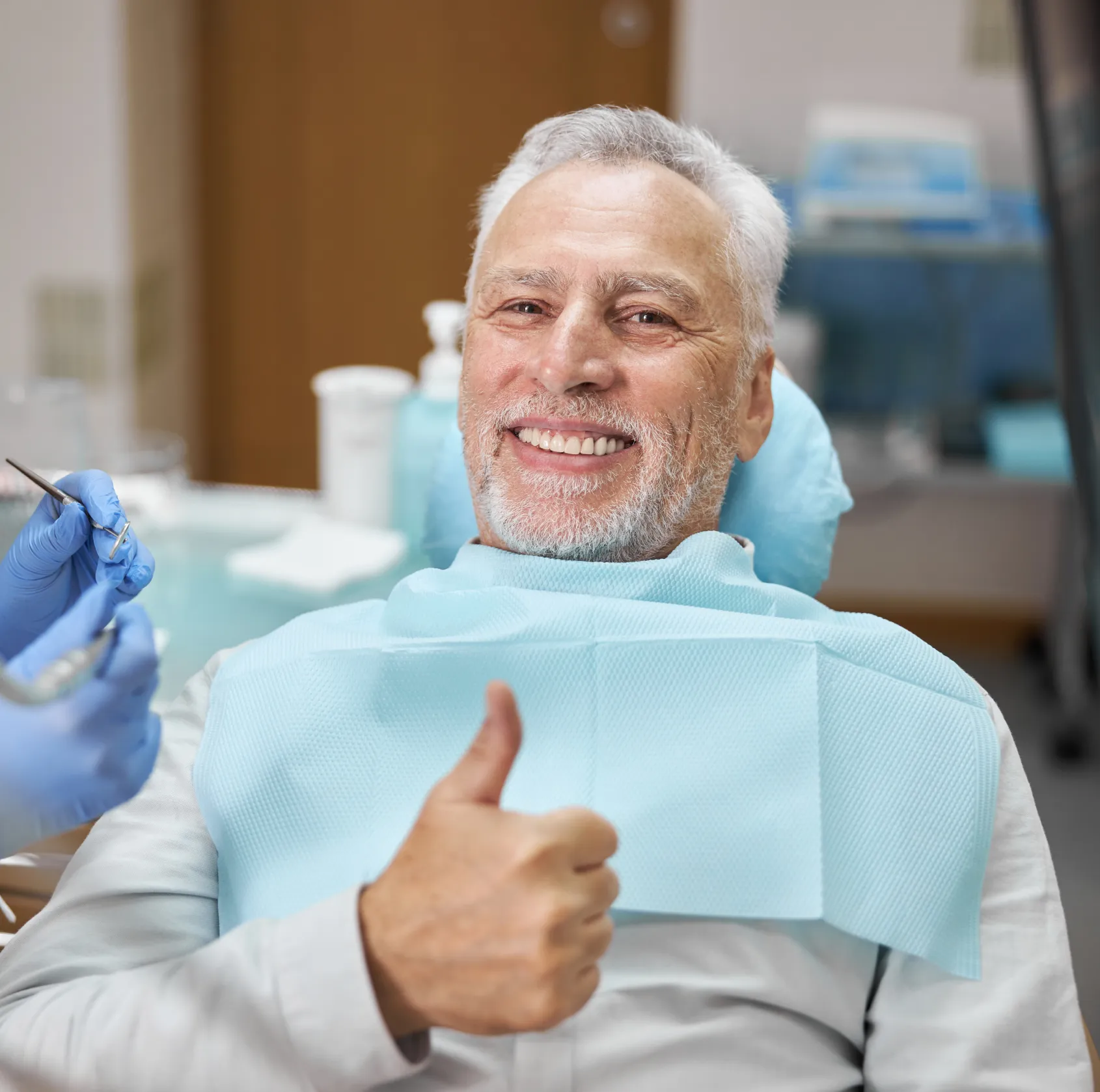 Smiling older man in dentist chair giving thumbs up with dental bib and tools nearby.
