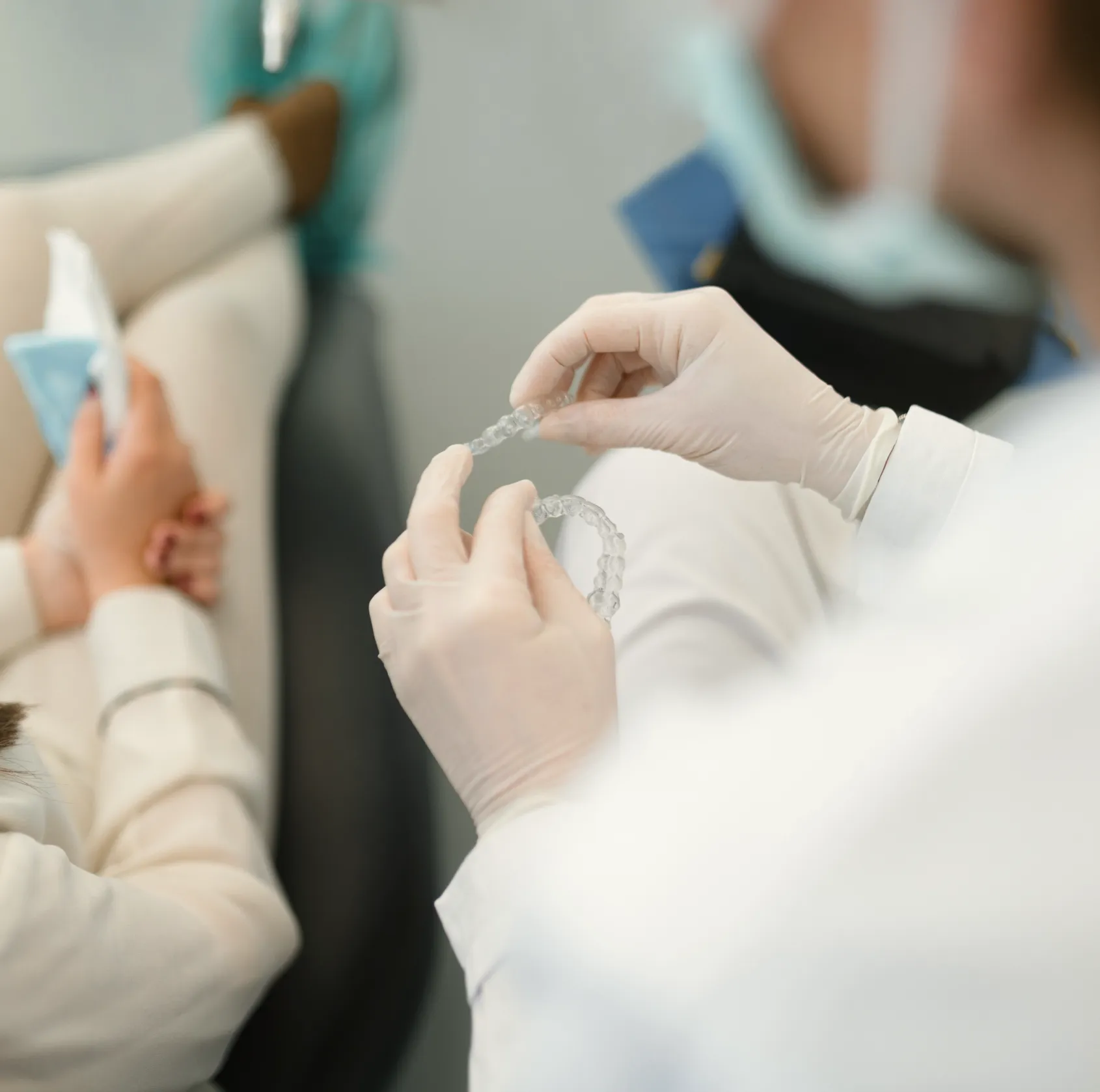 Dentist wearing gloves holding clear dental aligners while patient sits nearby in clinic.