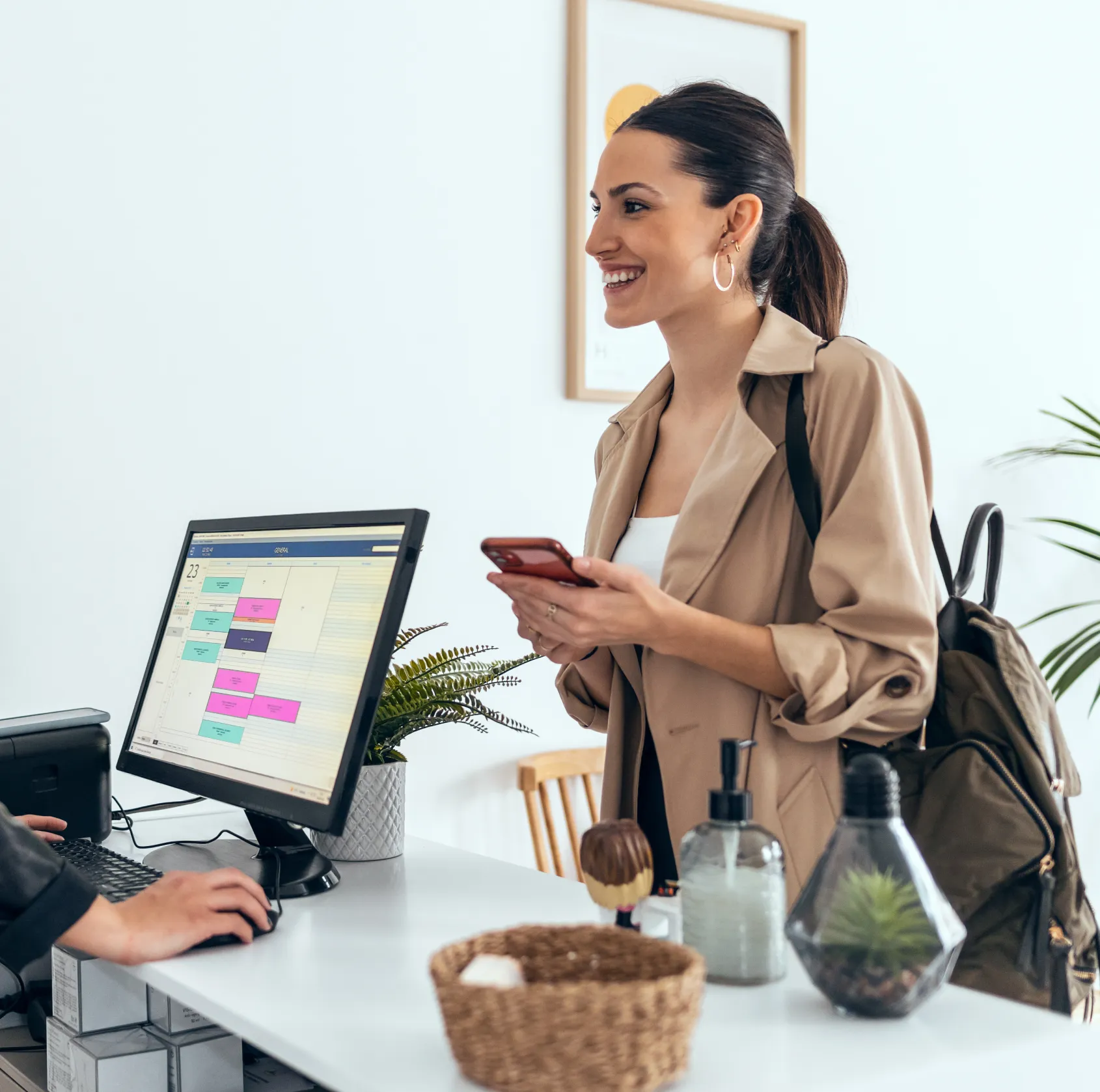 Smiling woman with backpack stands at reception desk holding phone as receptionist works on computer.