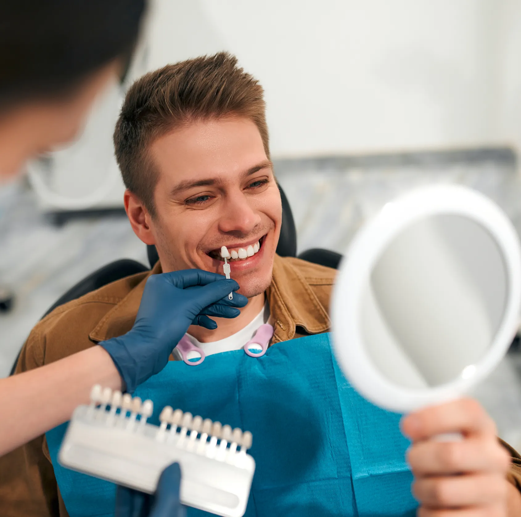 Dentist with blue gloves holds tooth shade guide to smiling man's teeth at dental clinic.