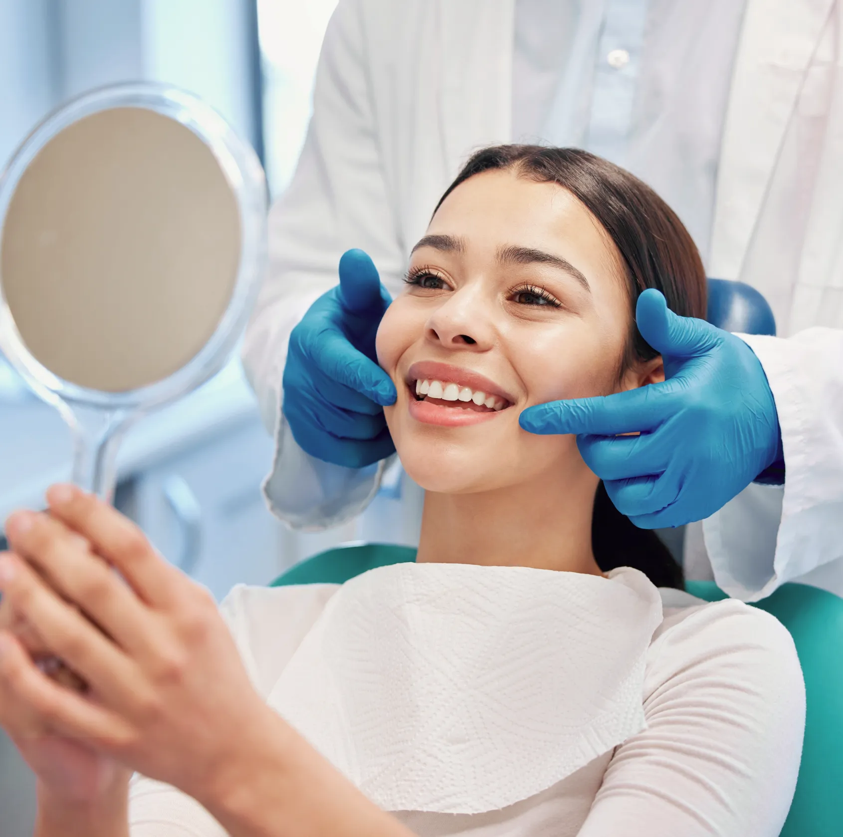 Dentist in blue gloves examines smiling woman's teeth as she looks in handheld mirror.