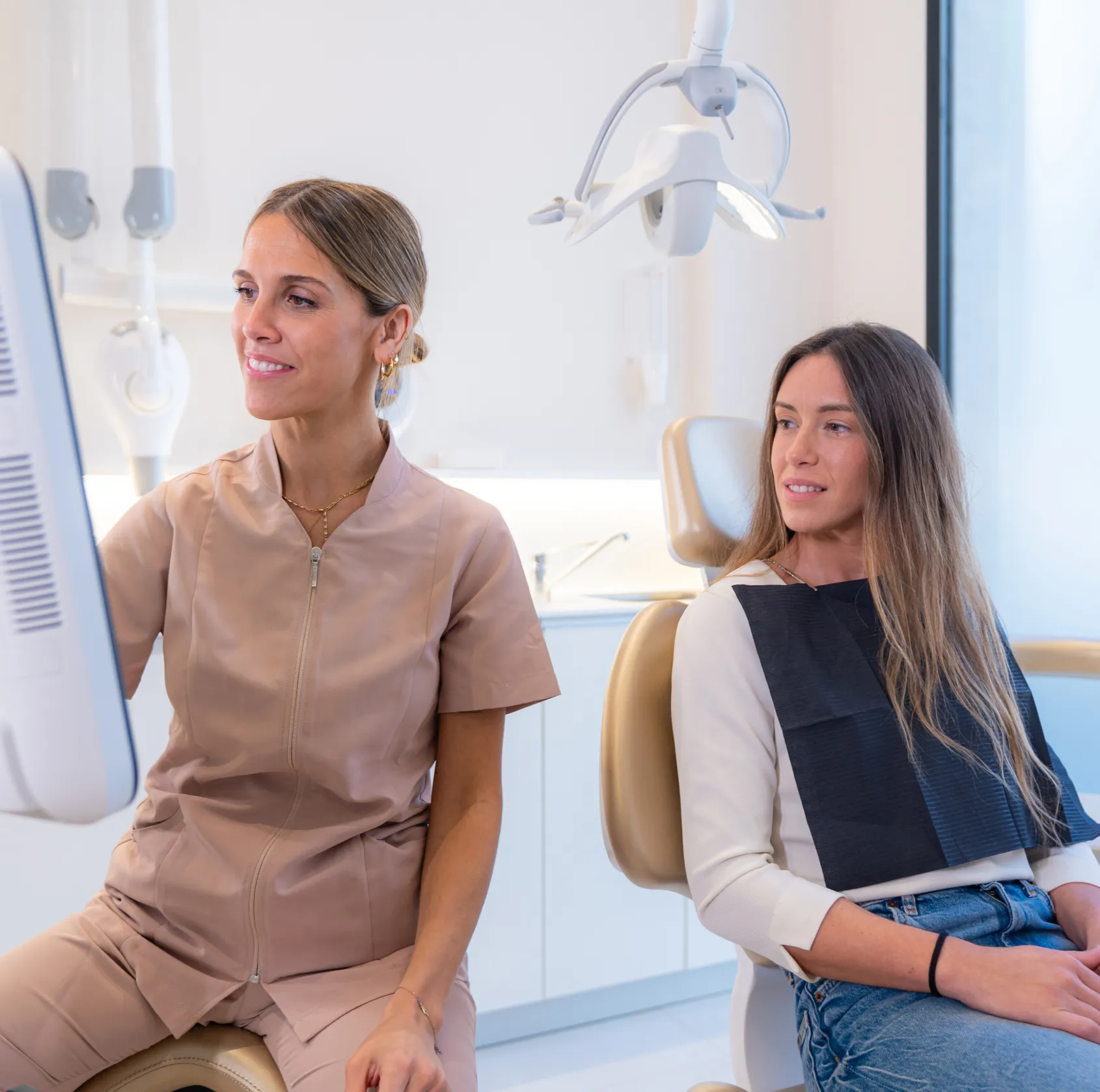 Dentist in beige scrubs reviewing a computer screen while female patient in a chair watches.