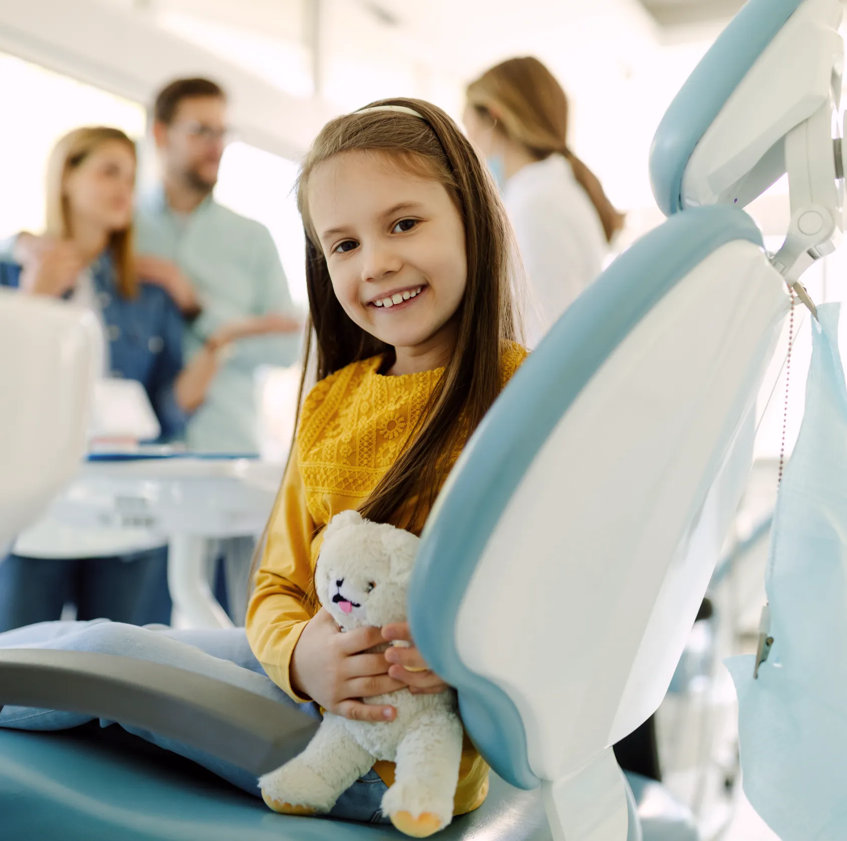 Smiling young girl holding a white teddy bear sitting in a dental chair with blurred adults in background.