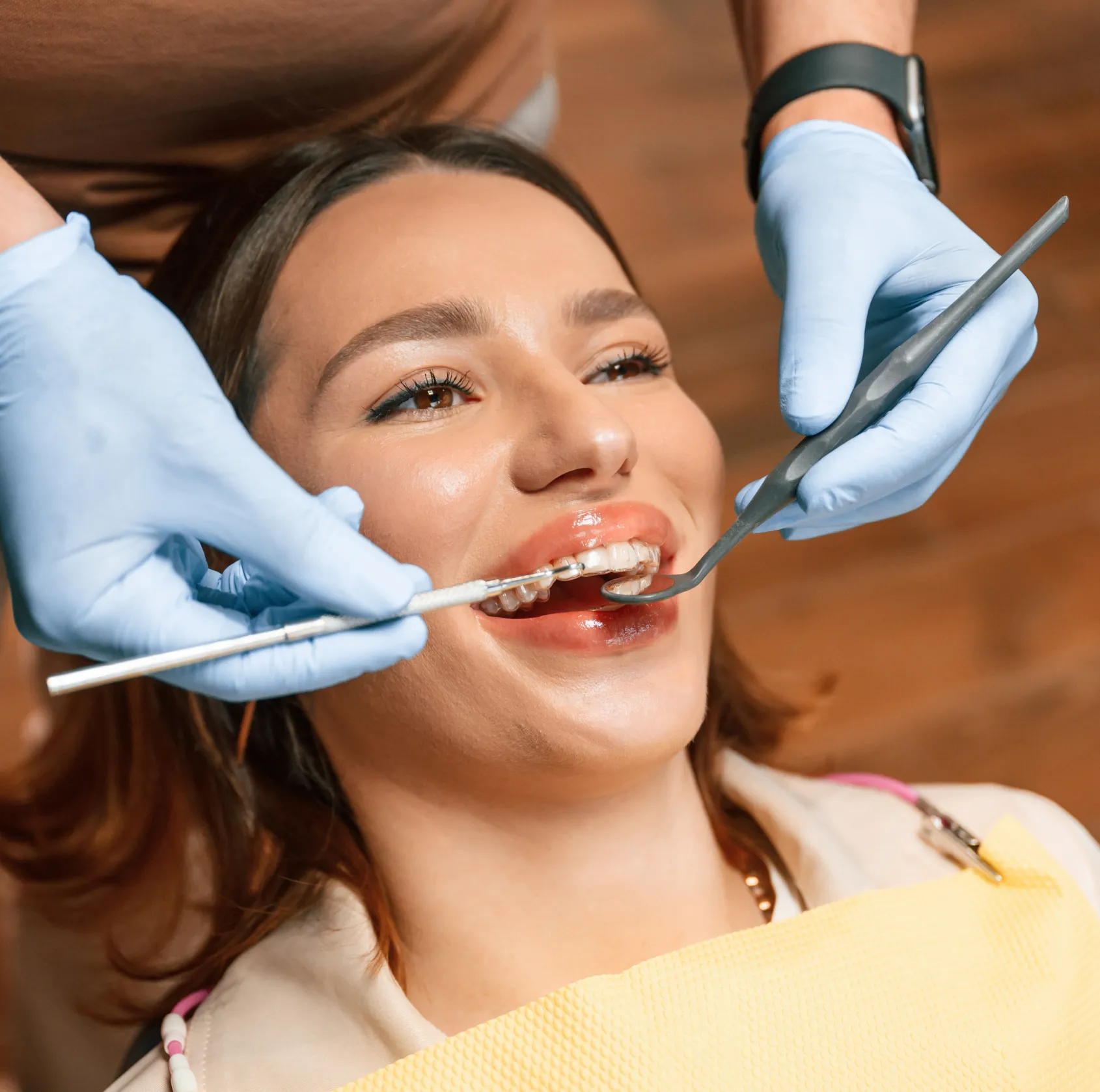 Dentist wearing gloves examines a smiling patient's teeth with dental tools and clear aligner.