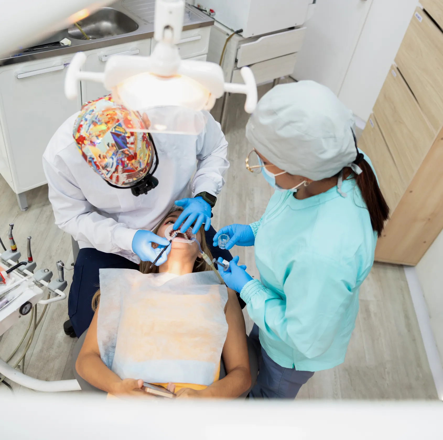 Dentist and assistant with gloves working on a patient lying in a dental chair under a dental light.