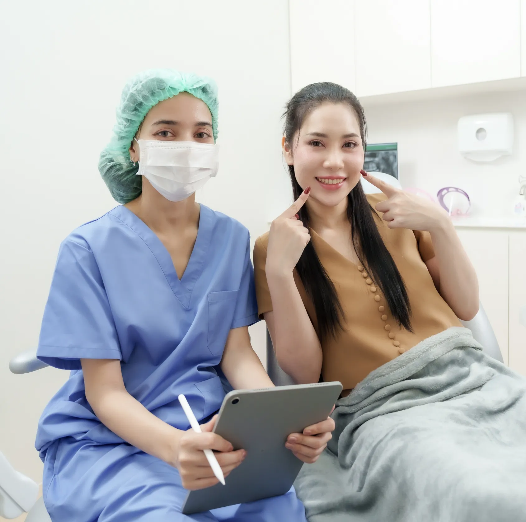 Healthcare worker in scrubs and mask with tablet next to smiling woman pointing at her cheeks.