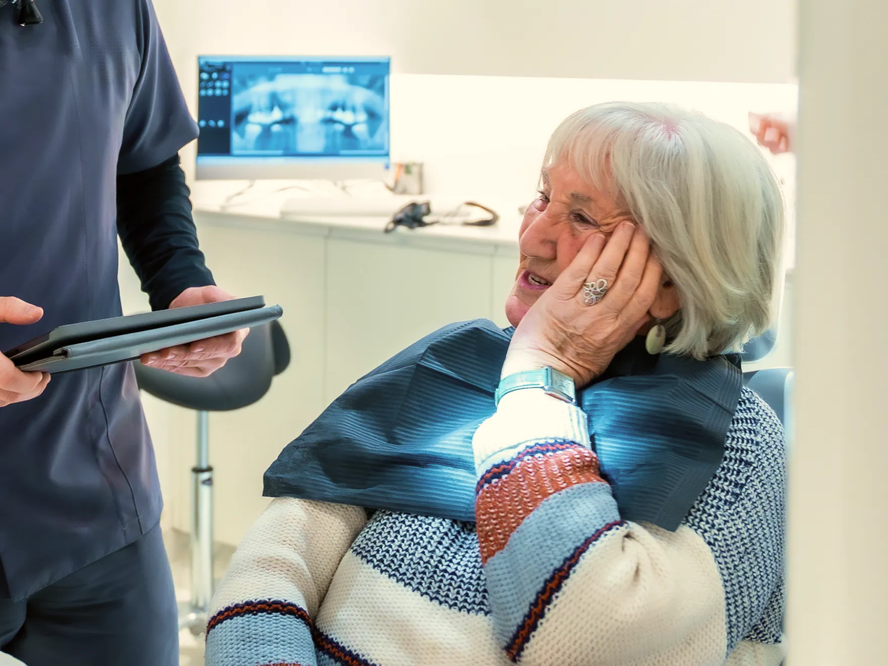 Elderly woman sitting in dental chair, holding her cheek, talking to a dental professional with tablet.