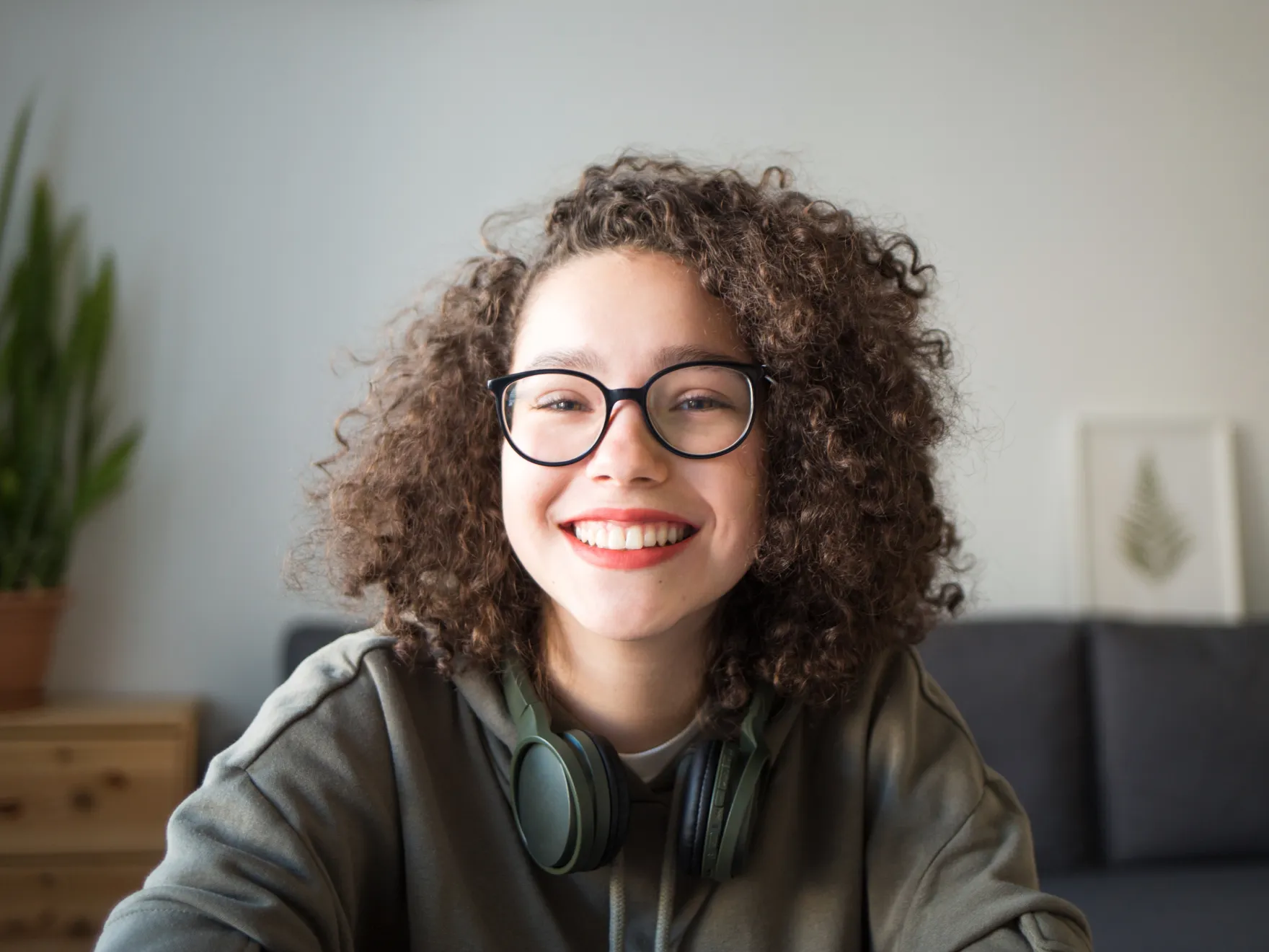 Smiling young woman with curly hair and glasses wearing a hoodie with headphones around her neck.