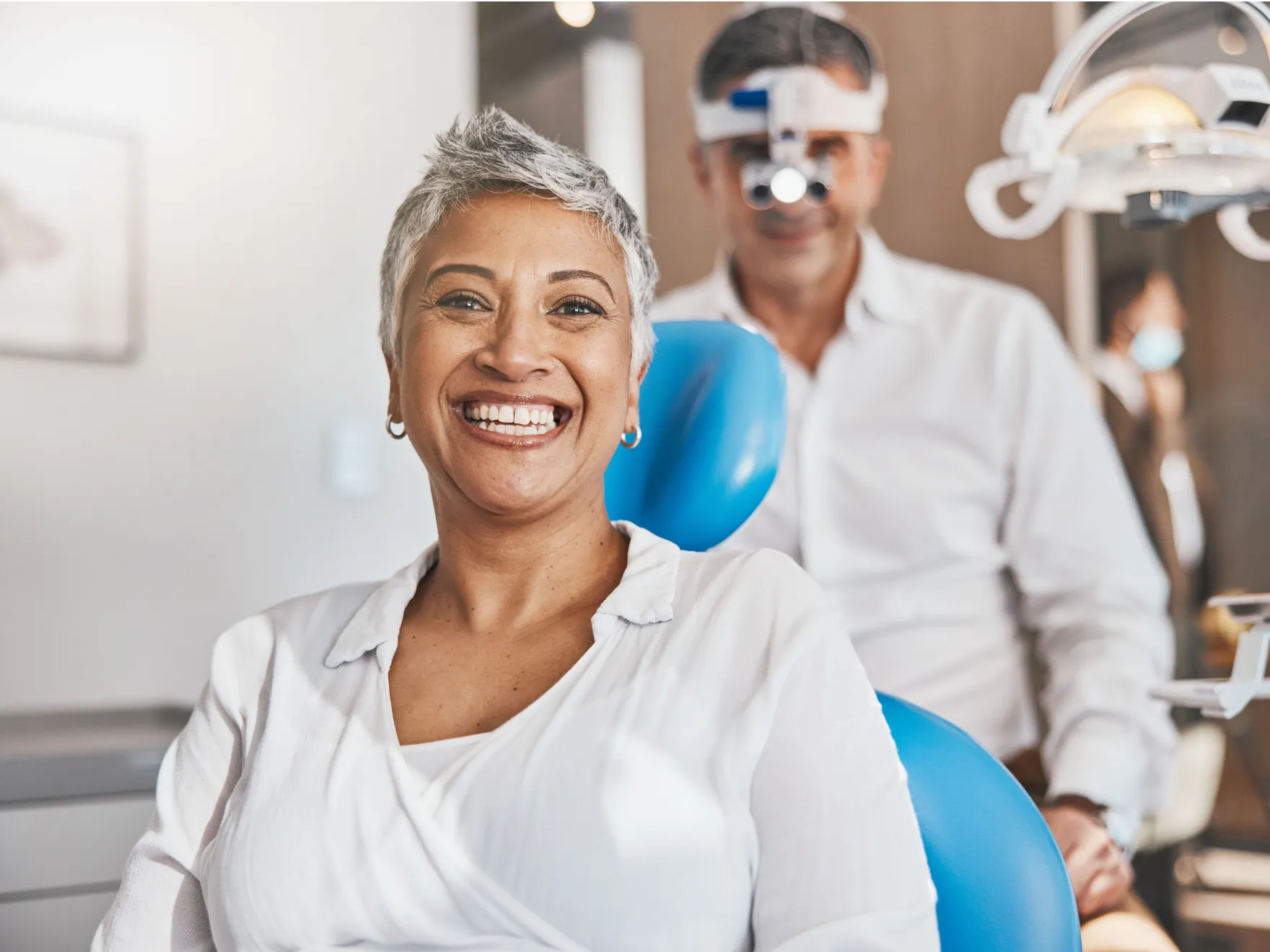 Smiling woman sitting in dental chair with dentist standing behind wearing magnifying glasses.