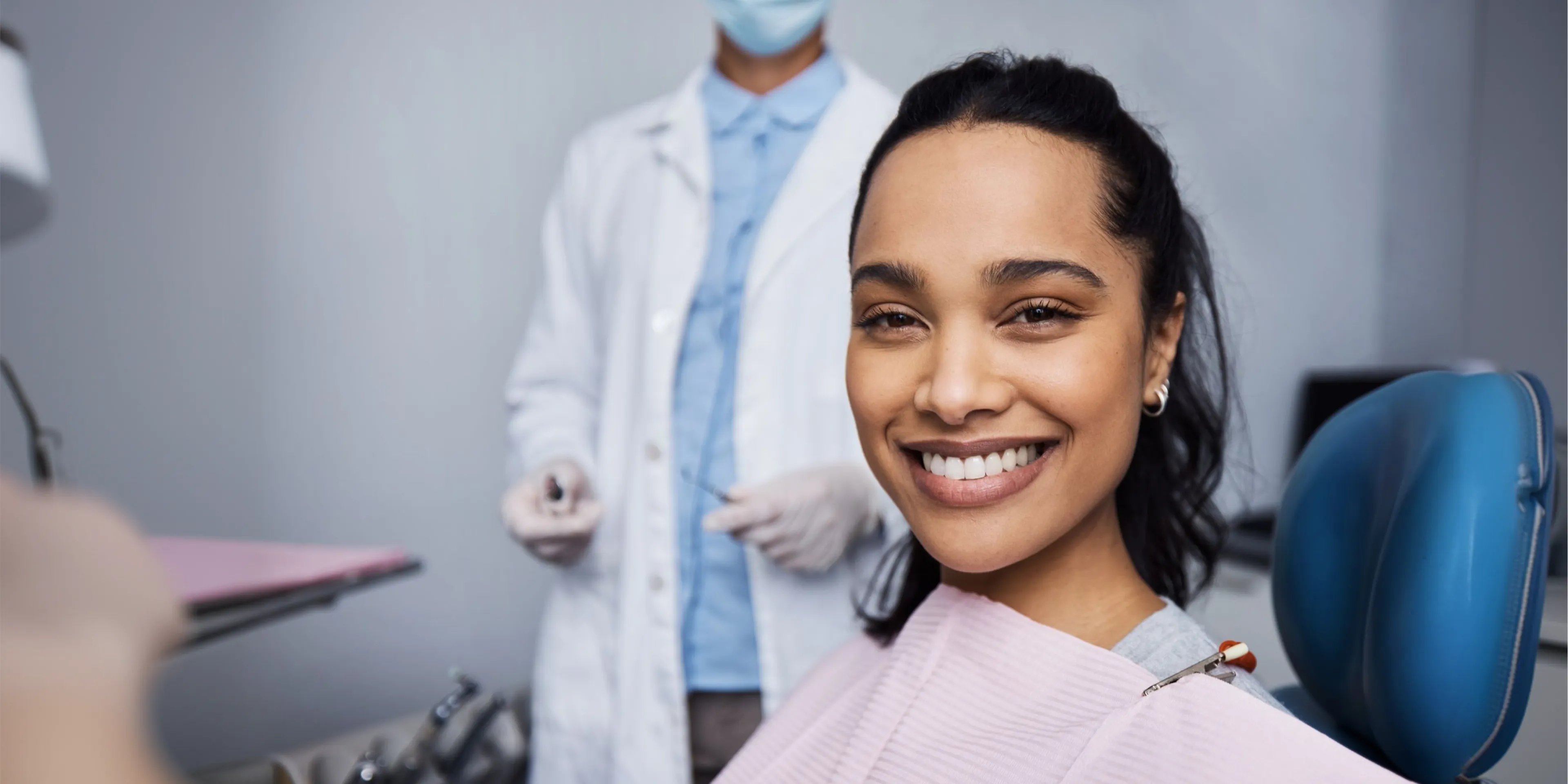 Smiling woman sitting in dental chair with dentist standing blurred in background.