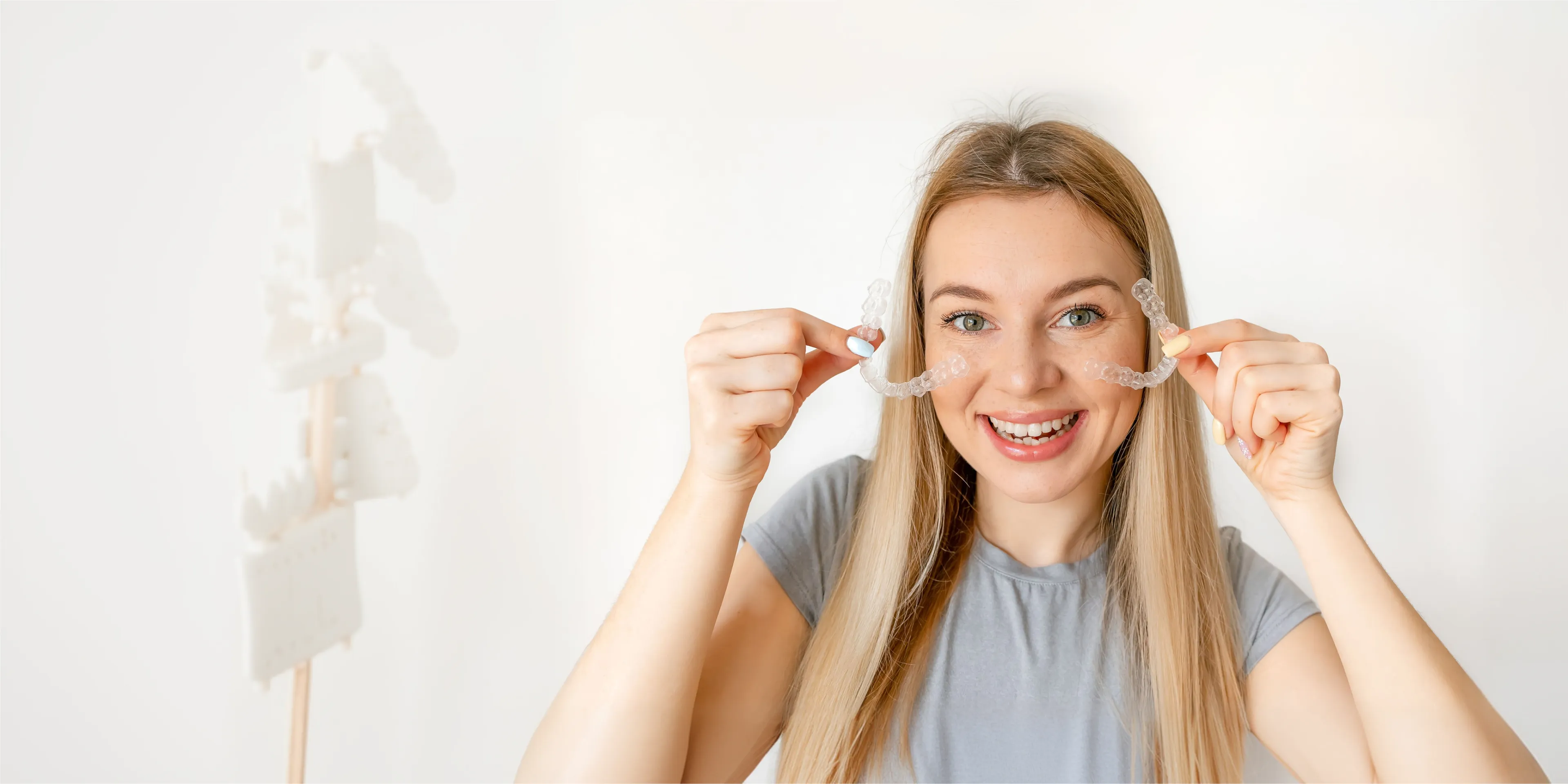 Smiling woman holding transparent dental aligners near her face on white background.