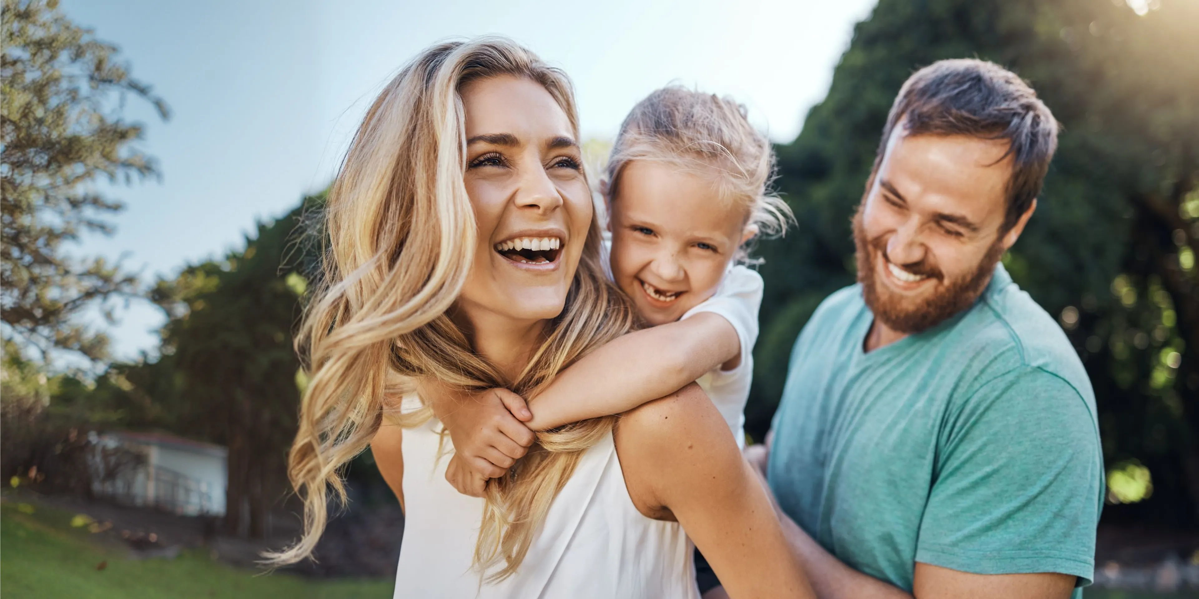 Happy family outdoors: smiling blonde woman with child on her back and bearded man in green shirt.
