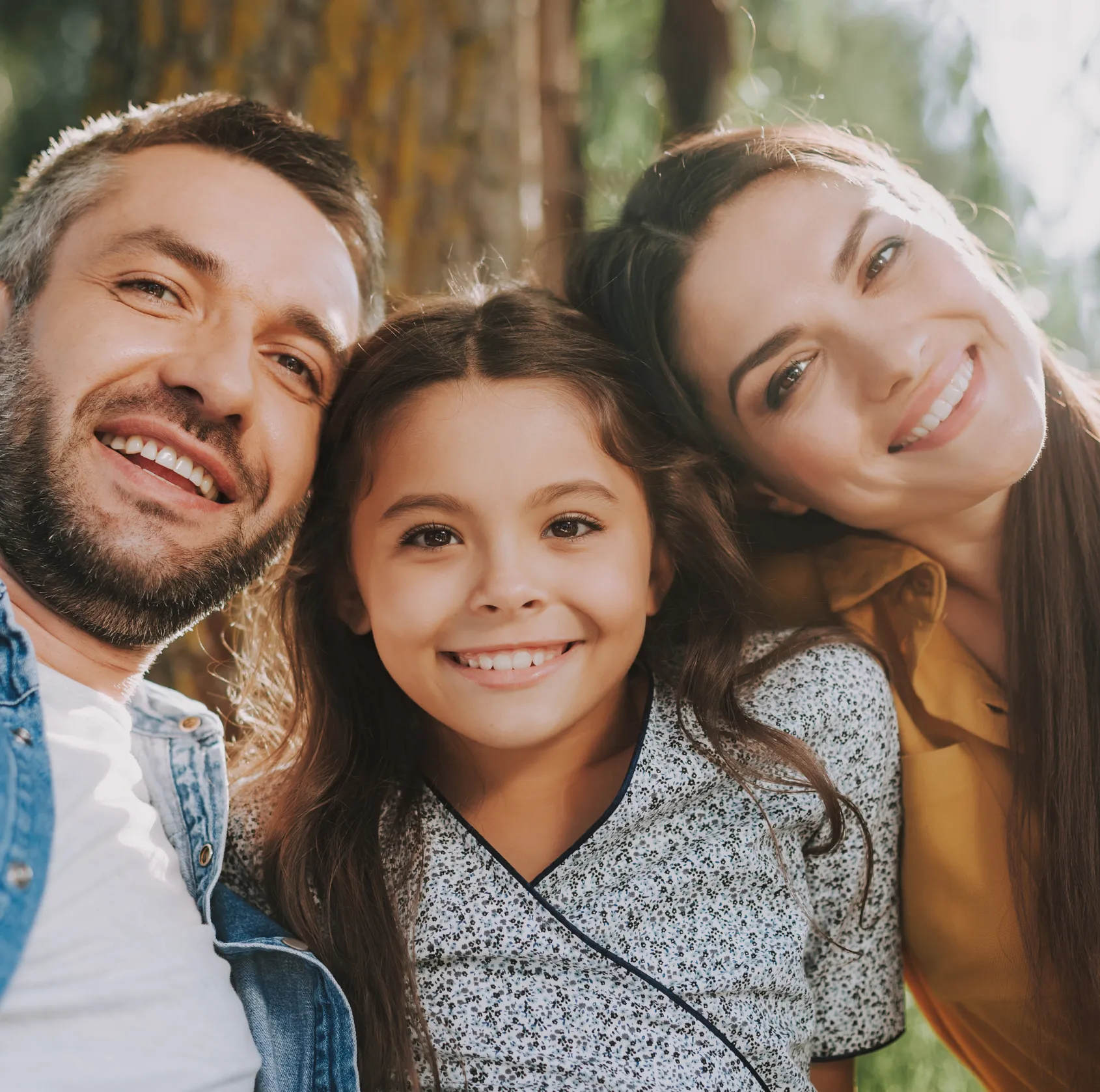 Smiling man, woman, and young girl posing outdoors together with trees in background.