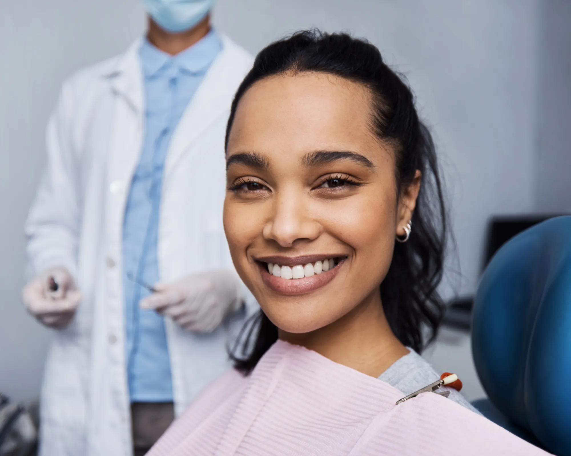 Smiling woman sitting in dental chair with dentist in mask and gloves in background.
