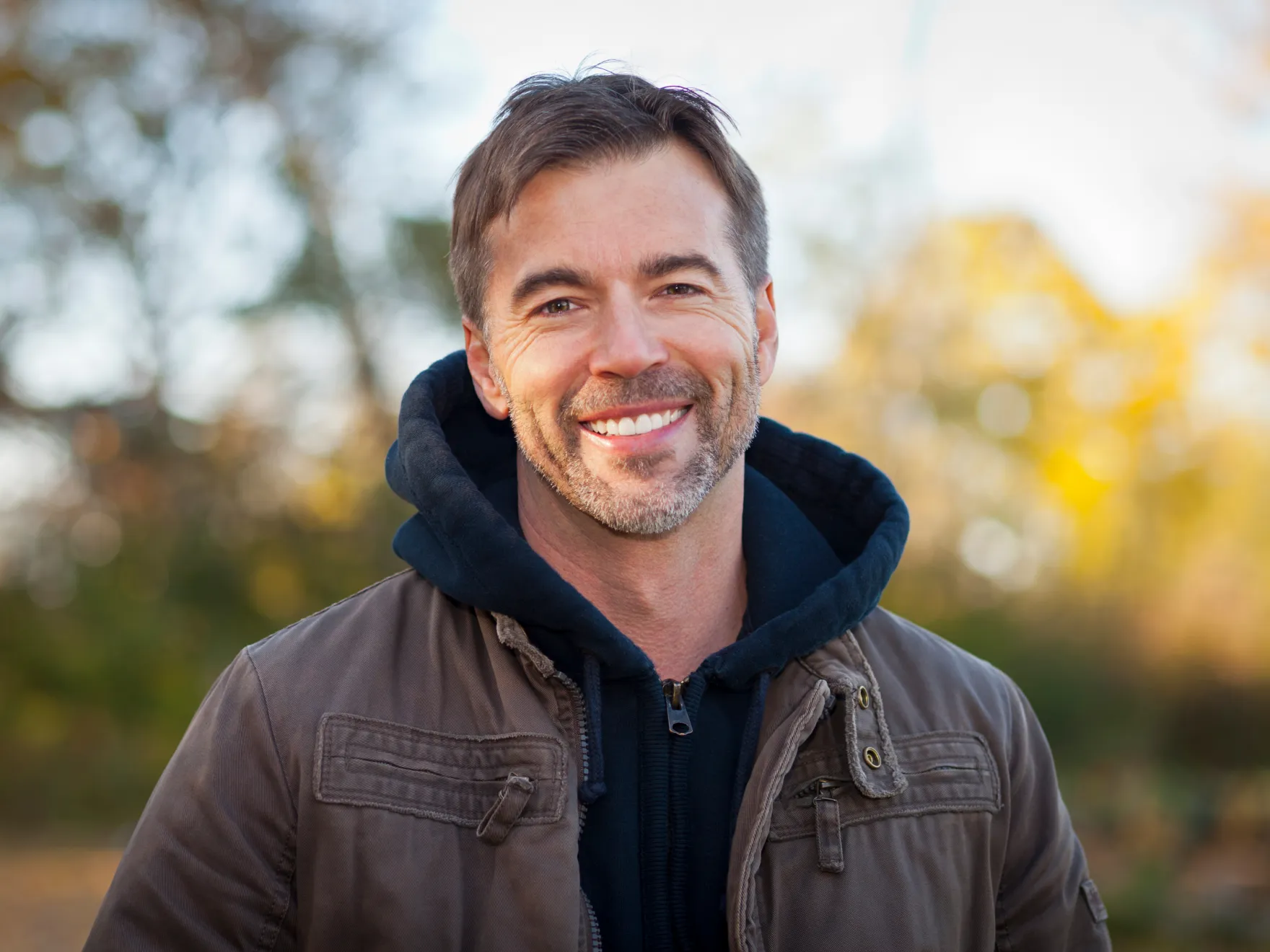 Smiling man with beard wearing a brown jacket and black hoodie outdoors in autumn.