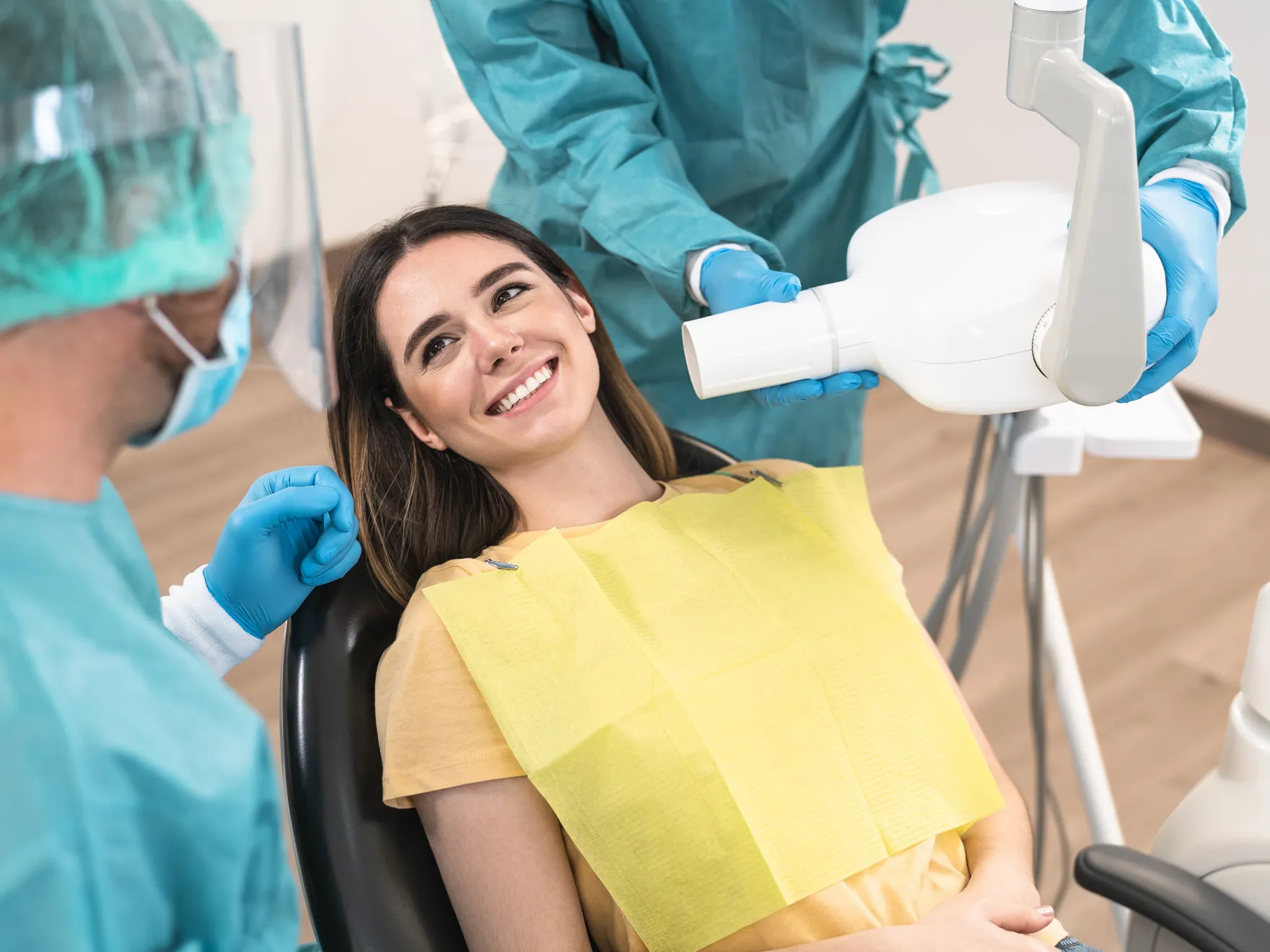 Smiling woman in dental chair with dental bib, dentist and assistant preparing x-ray machine.