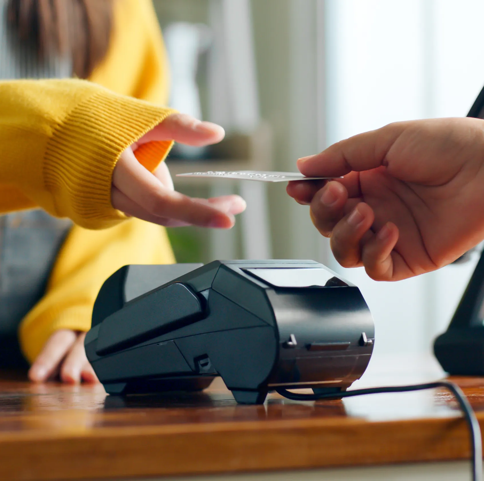 Person in yellow sweater reaching to receive a credit card from another hand over a card reader terminal.