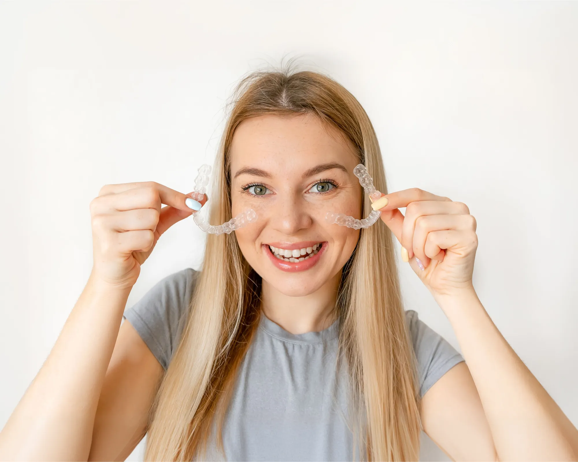 Smiling woman holding clear dental aligners near her face with a white background.