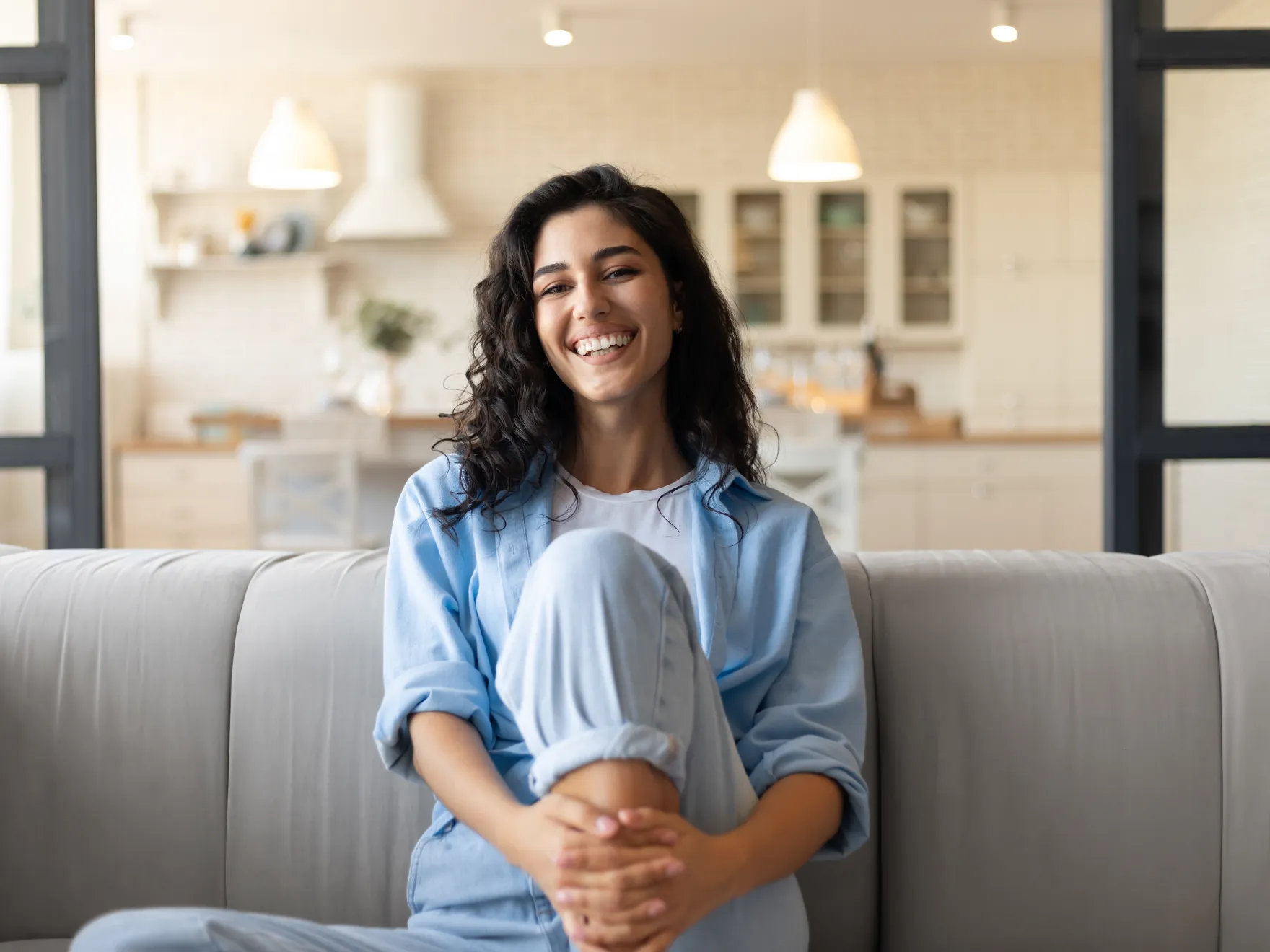 Smiling woman with curly hair sitting cross-legged on a couch in a bright living room.