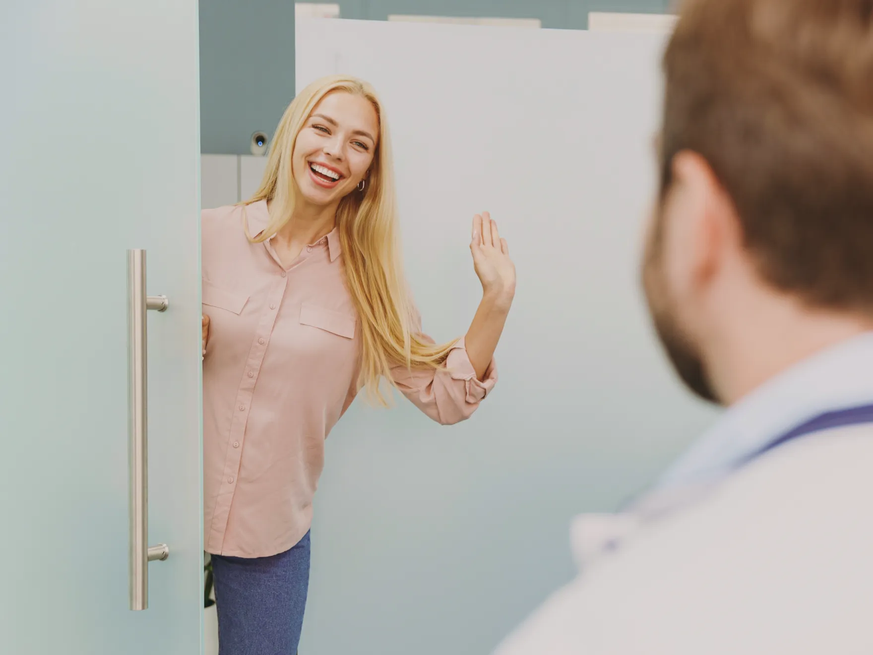 Smiling woman waving while standing in a doorway, facing a man with a stethoscope.