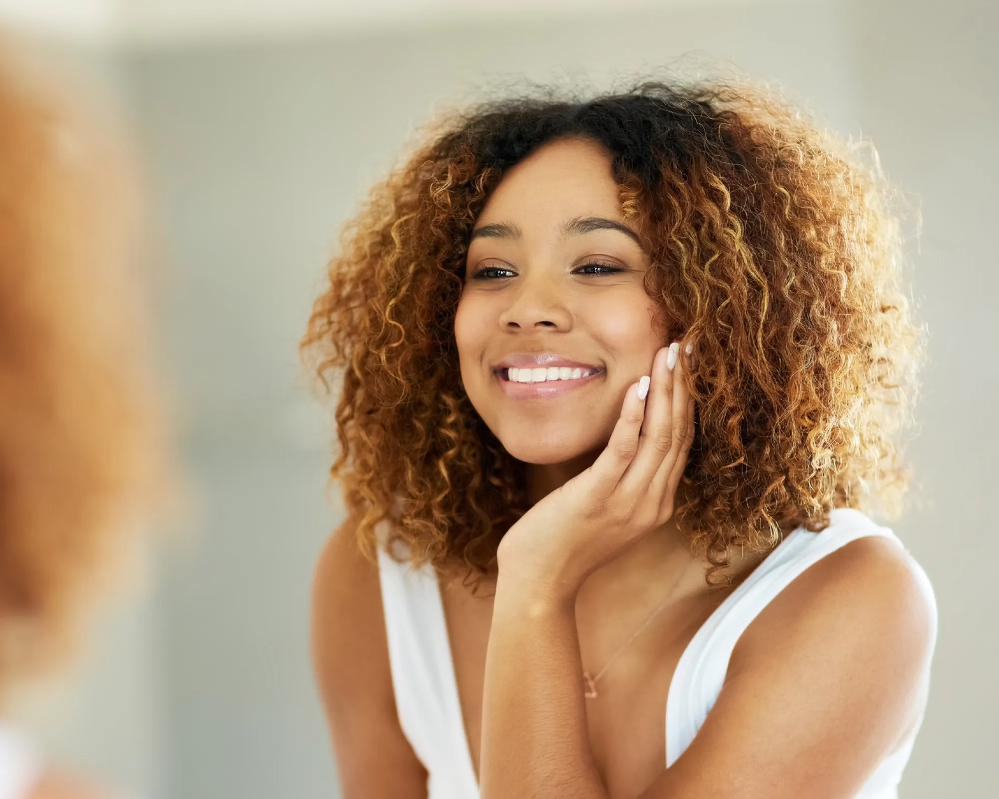 Smiling woman with curly hair touching her face and looking in a mirror.
