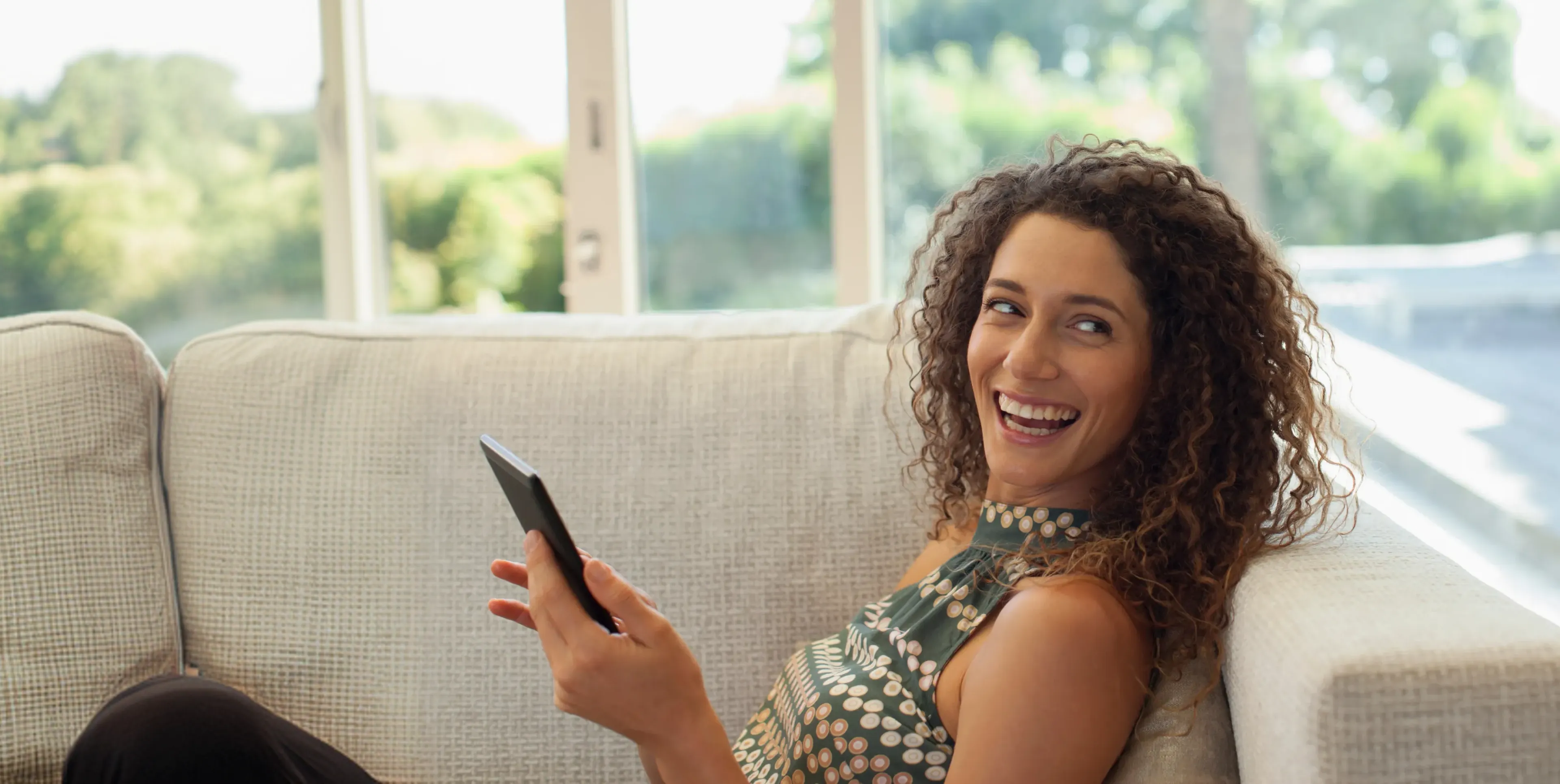 Smiling woman with curly hair holding a smartphone while sitting on a beige couch near a window.