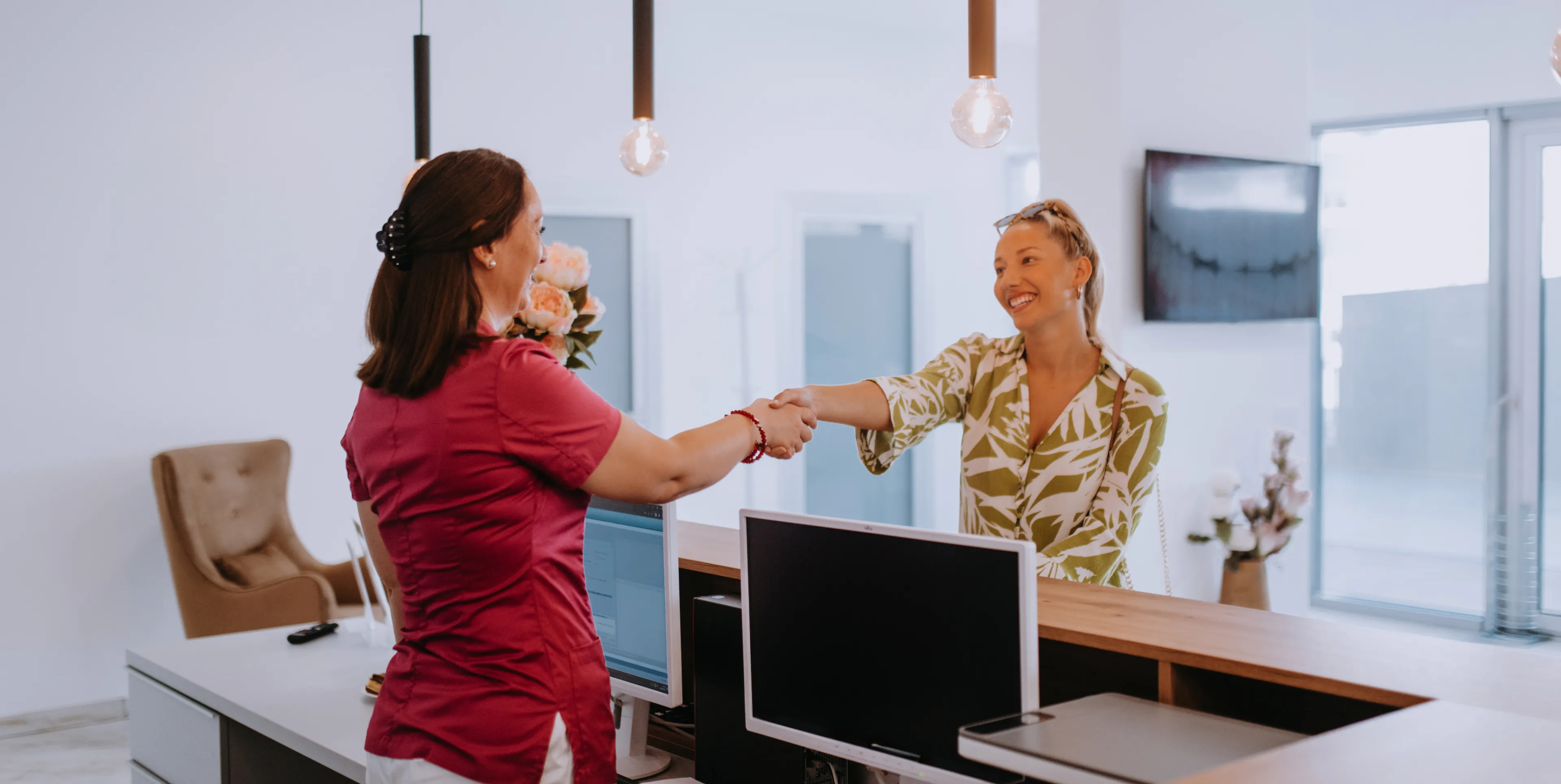 Receptionist in red shirt shaking hands with smiling woman in green leaf print blouse at desk.