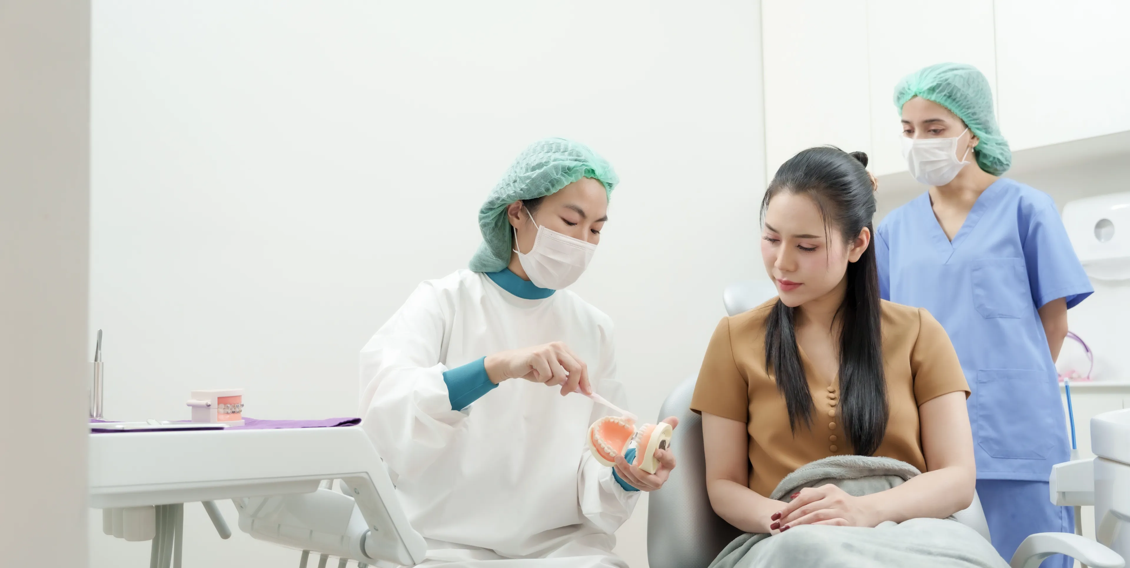 Dentist in protective wear showing a dental model and brushing technique to a seated woman patient.
