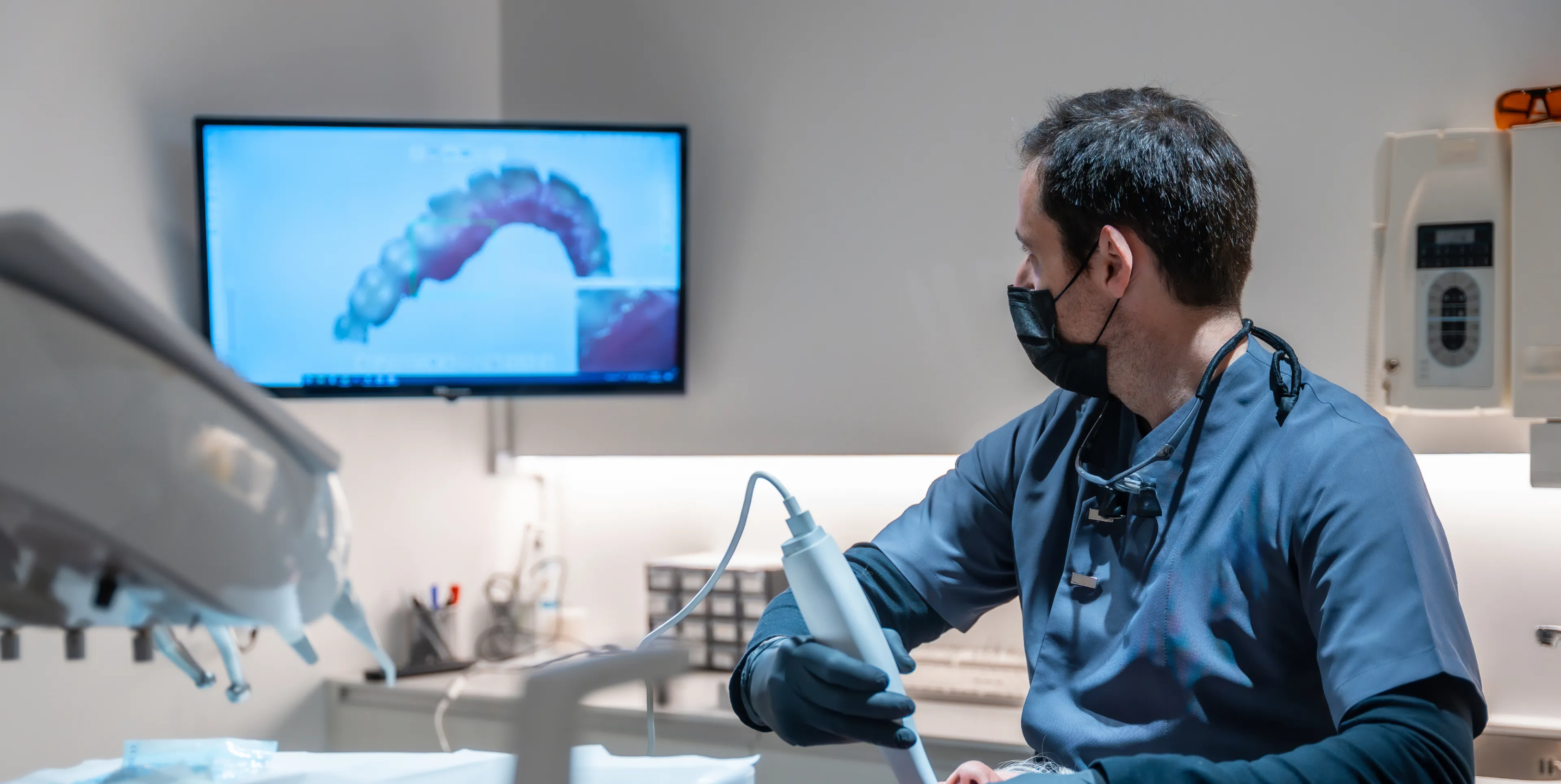 Dentist in mask using digital scanner to create 3D dental impression displayed on screen.