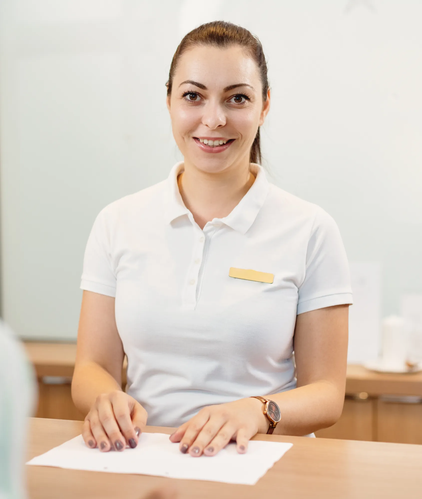 Smiling woman in a white polo shirt with a blank name tag sitting at a table with papers.