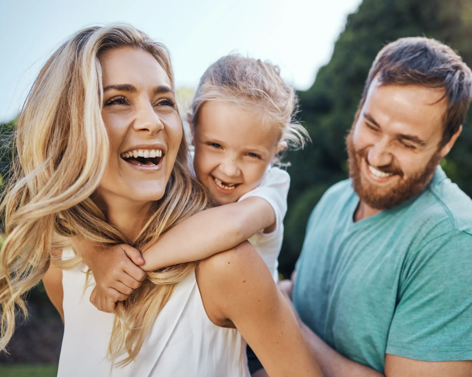 Smiling child hugs woman from behind while man looks on happily outdoors.