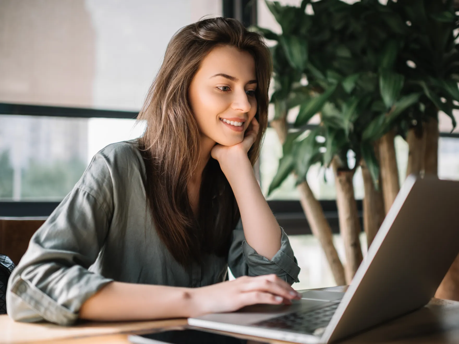 Smiling woman with long hair using laptop at a desk with a large plant in the background.