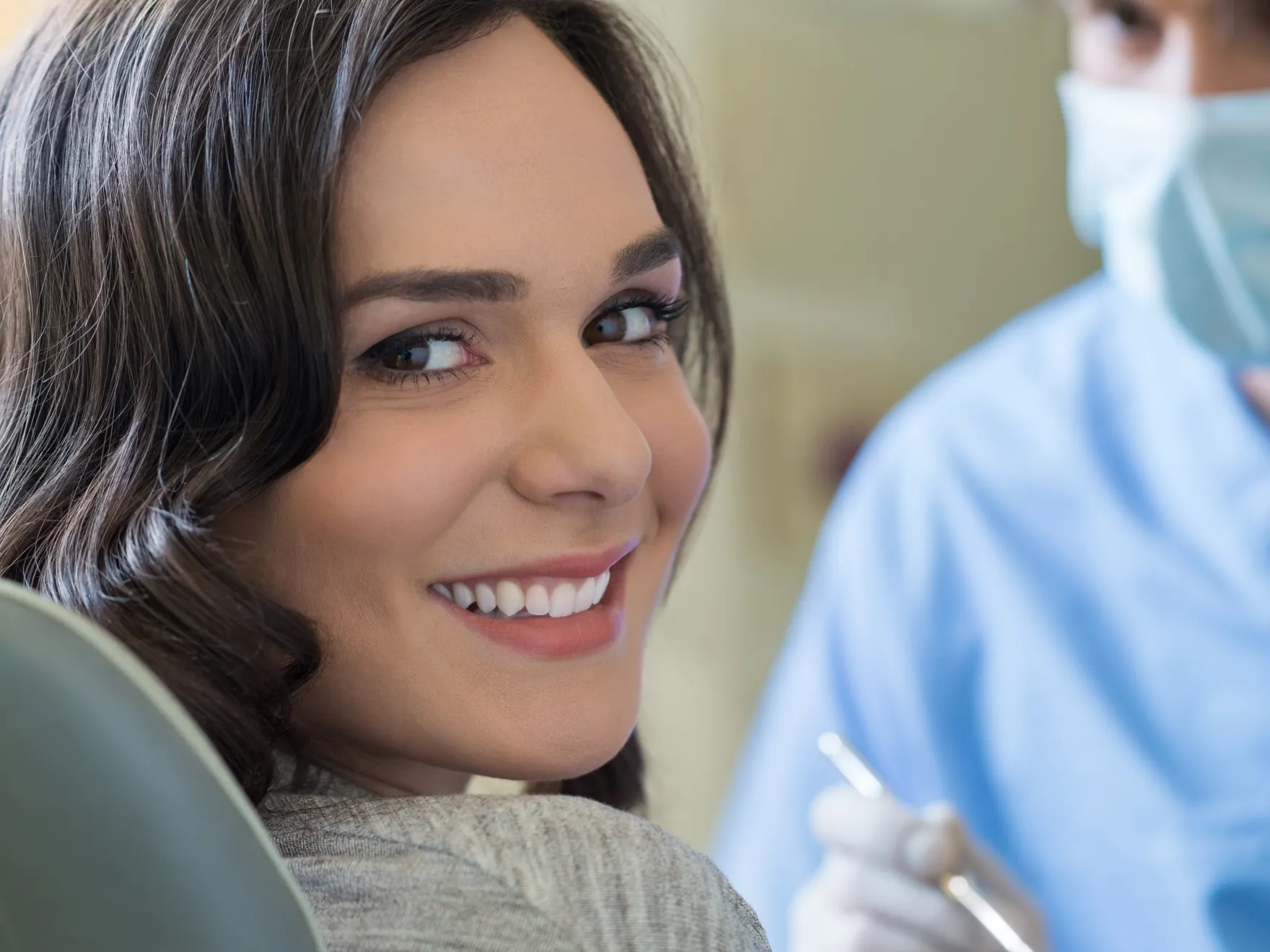 Smiling woman sitting in a dental chair with a masked dentist holding a tool in the background.