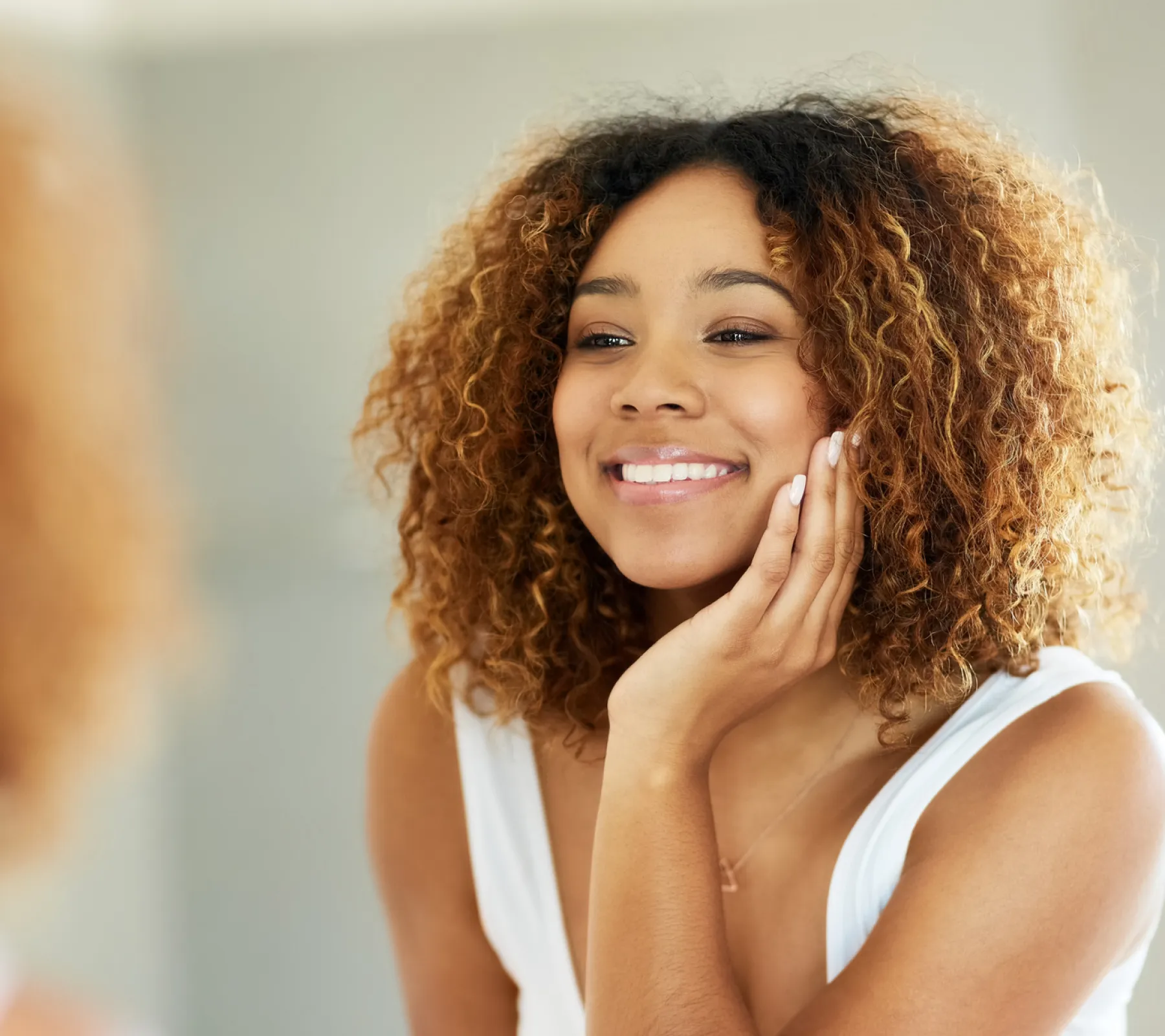 Smiling woman with curly hair touching her face while looking in a mirror.