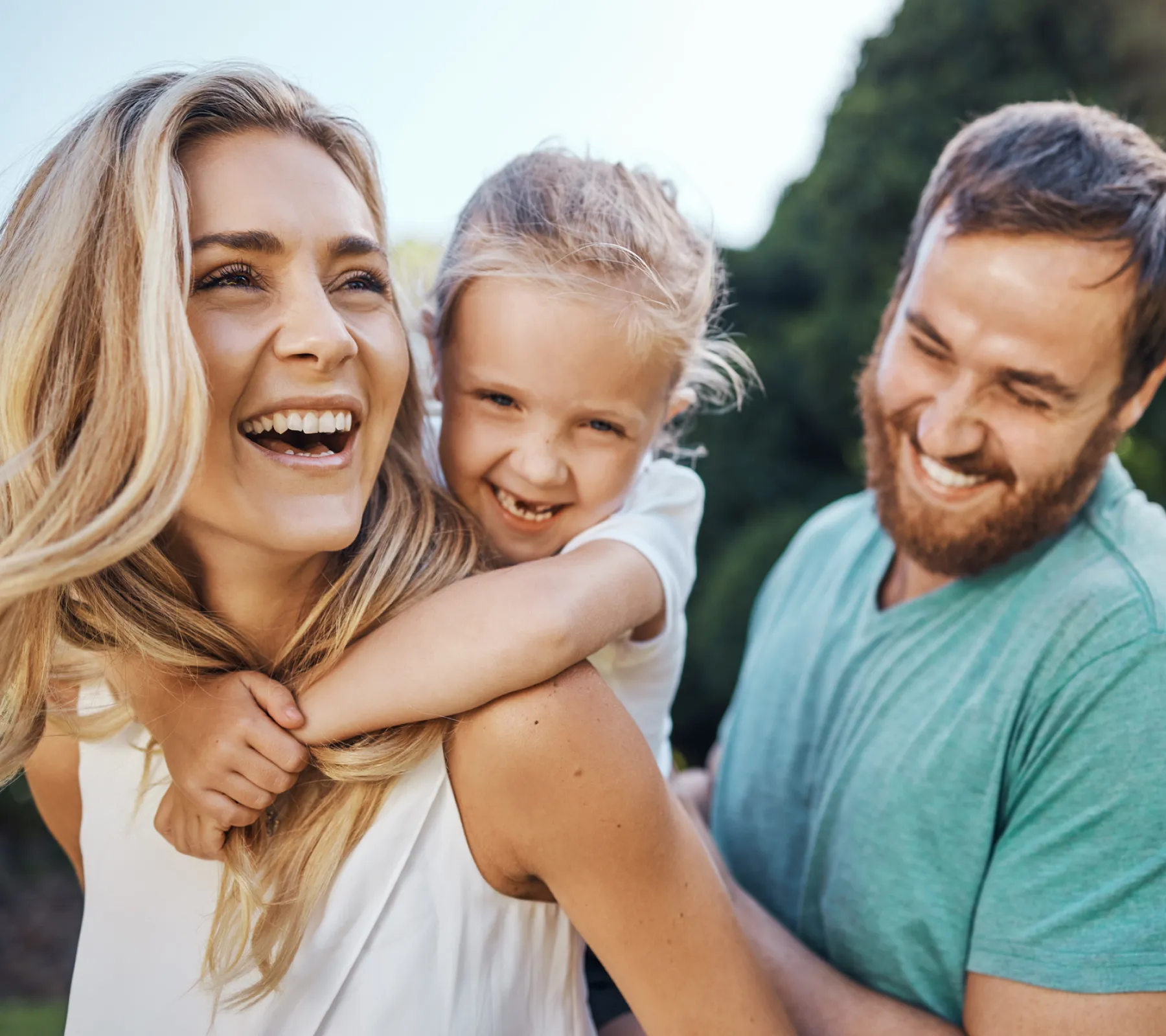 Smiling family outdoors: woman carrying child on back and man laughing beside them.