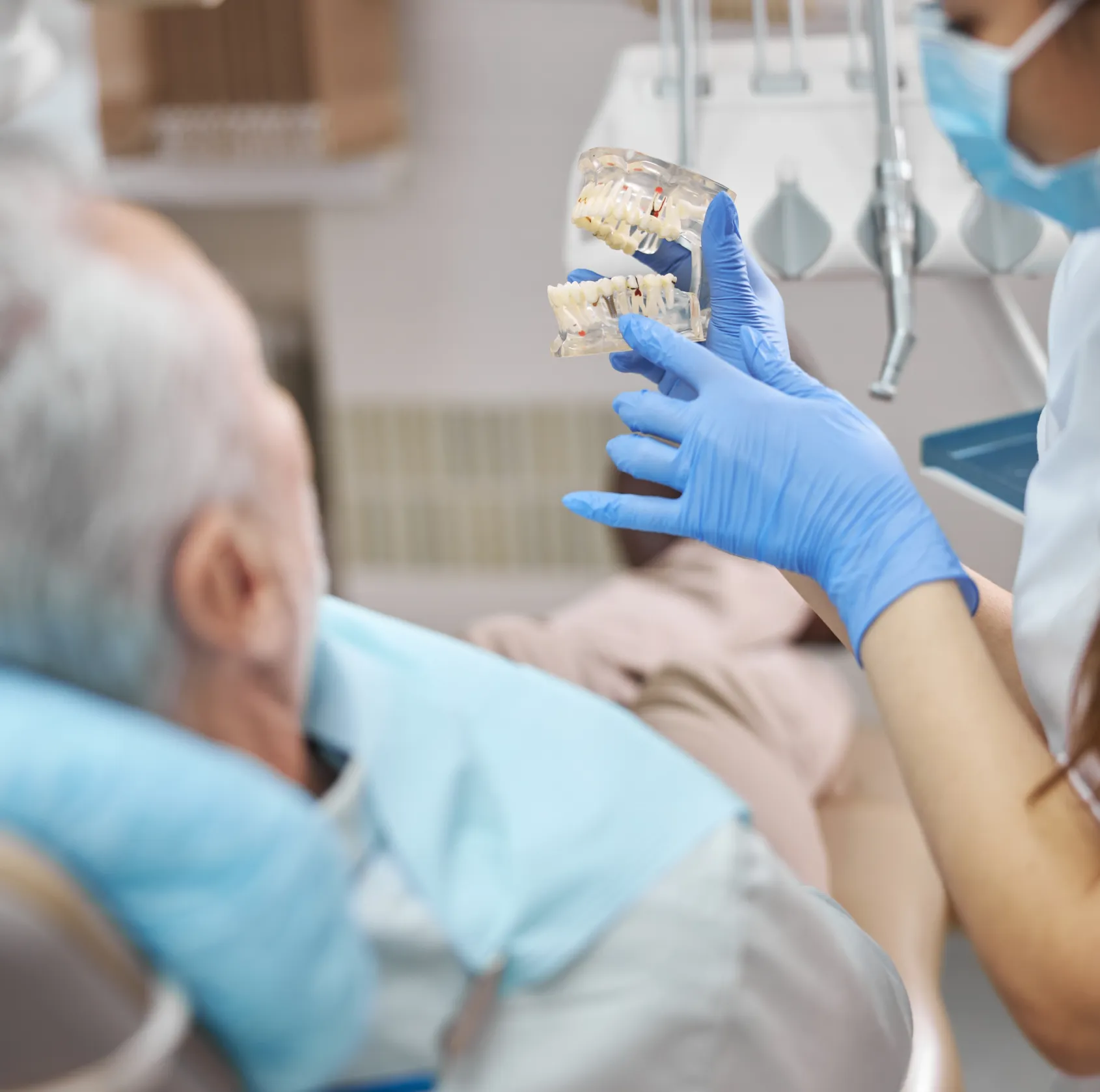 Dentist wearing blue gloves holds a clear dental jaw model showing teeth, explaining something to an older male patient reclining in a dental chair.