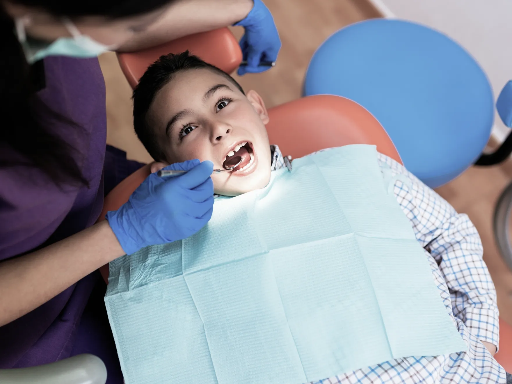 Young boy reclined in a dental chair with mouth open as a dental professional wearing blue gloves uses a dental mirror to examine his teeth, while the boy wears a protective bib.