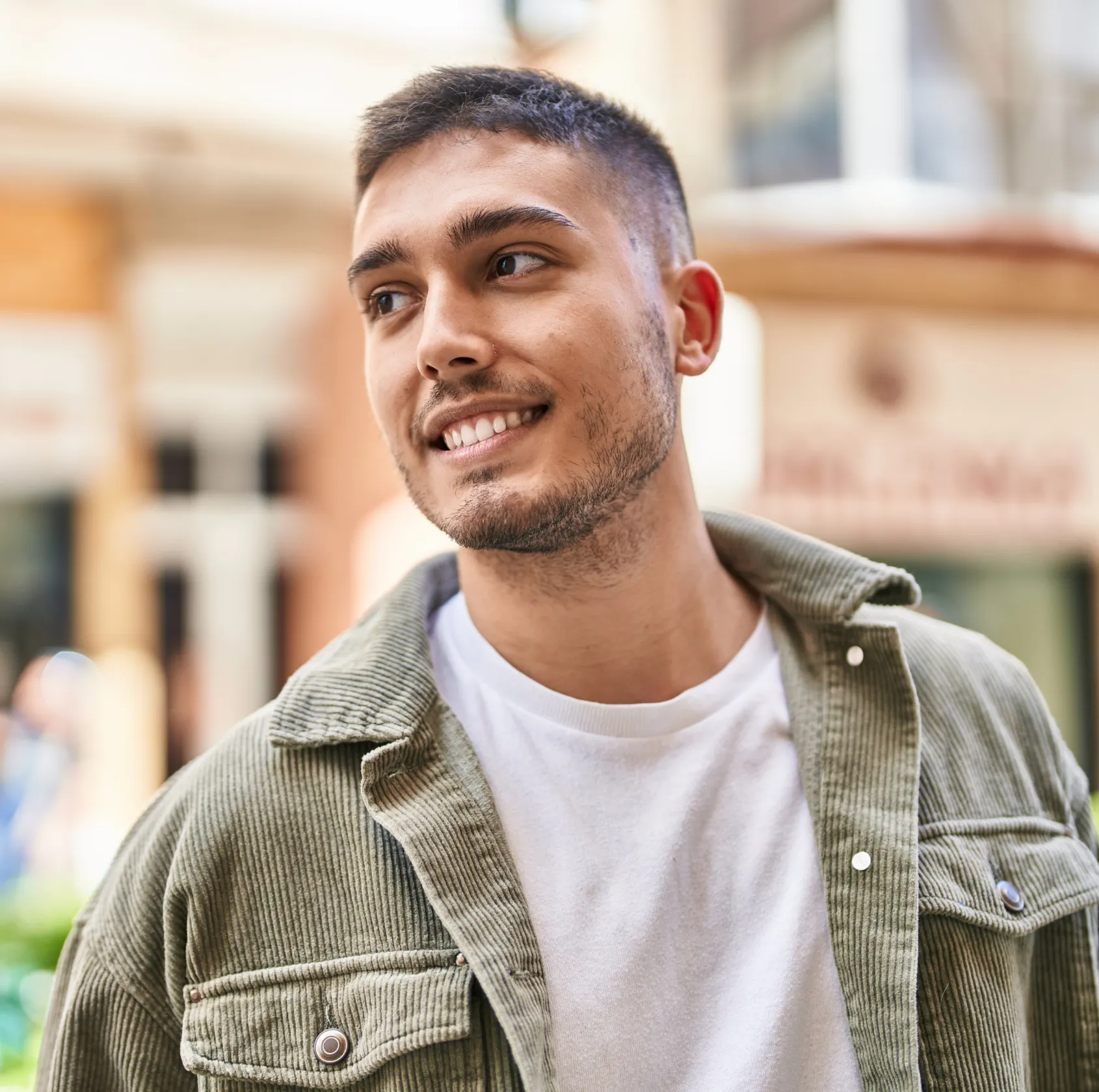 Young man with short dark hair and light stubble wearing a white T-shirt and green corduroy jacket smiling and looking to the side outdoors in an urban setting.