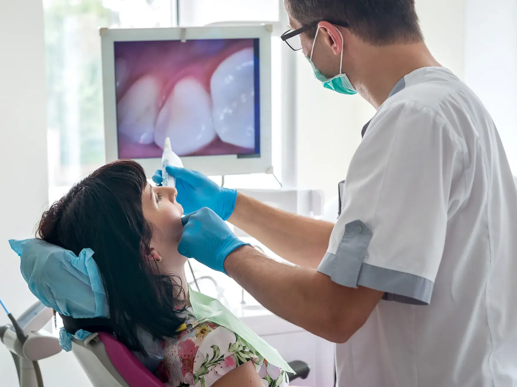 Dentist wearing mask and gloves examining a female patient's teeth using an intraoral camera, with a close-up image of teeth displayed on a monitor in the background.