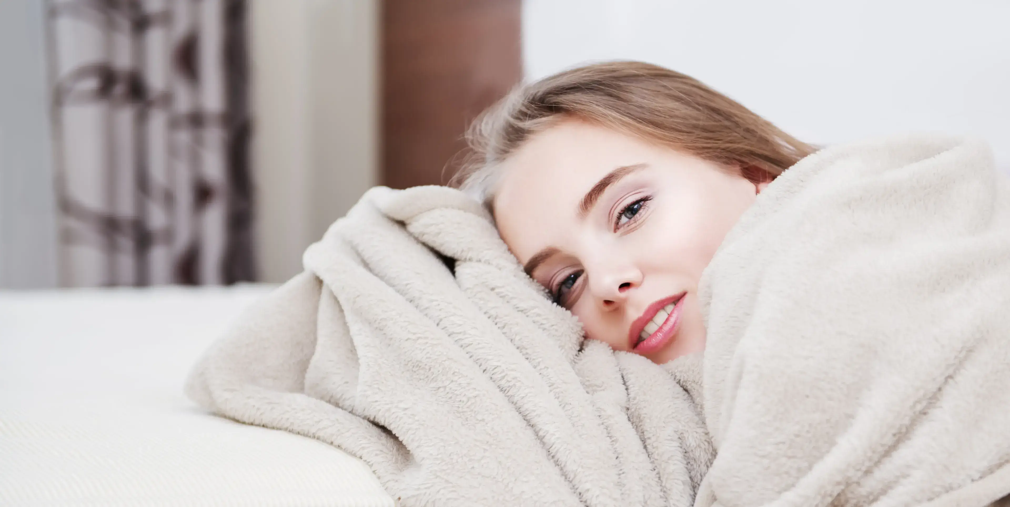 Close-up of a young woman lying down wrapped in a soft beige blanket, smiling gently at the camera.