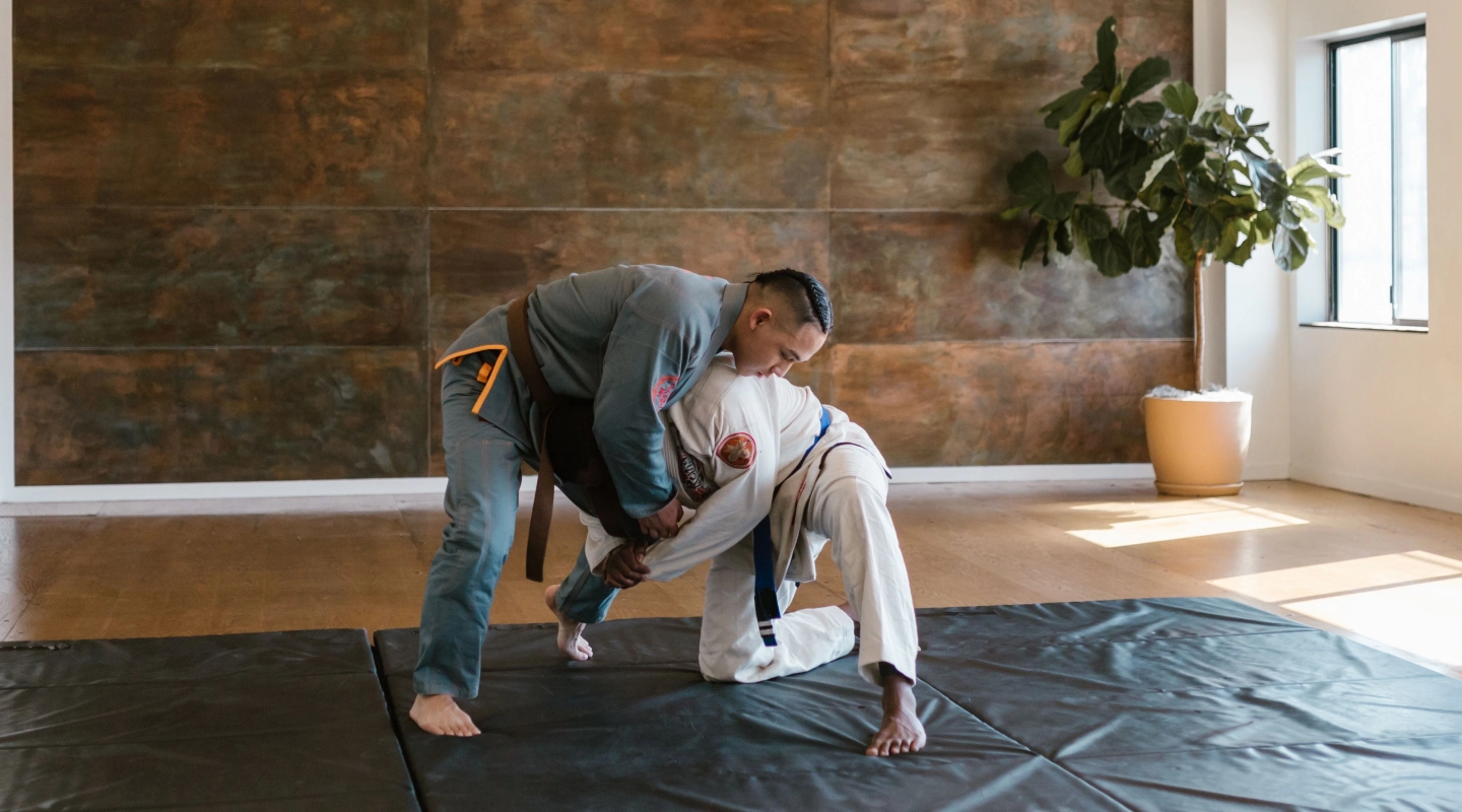 Two men engaged in martial arts training on a black mat in a room with a brick wall and a potted plant.