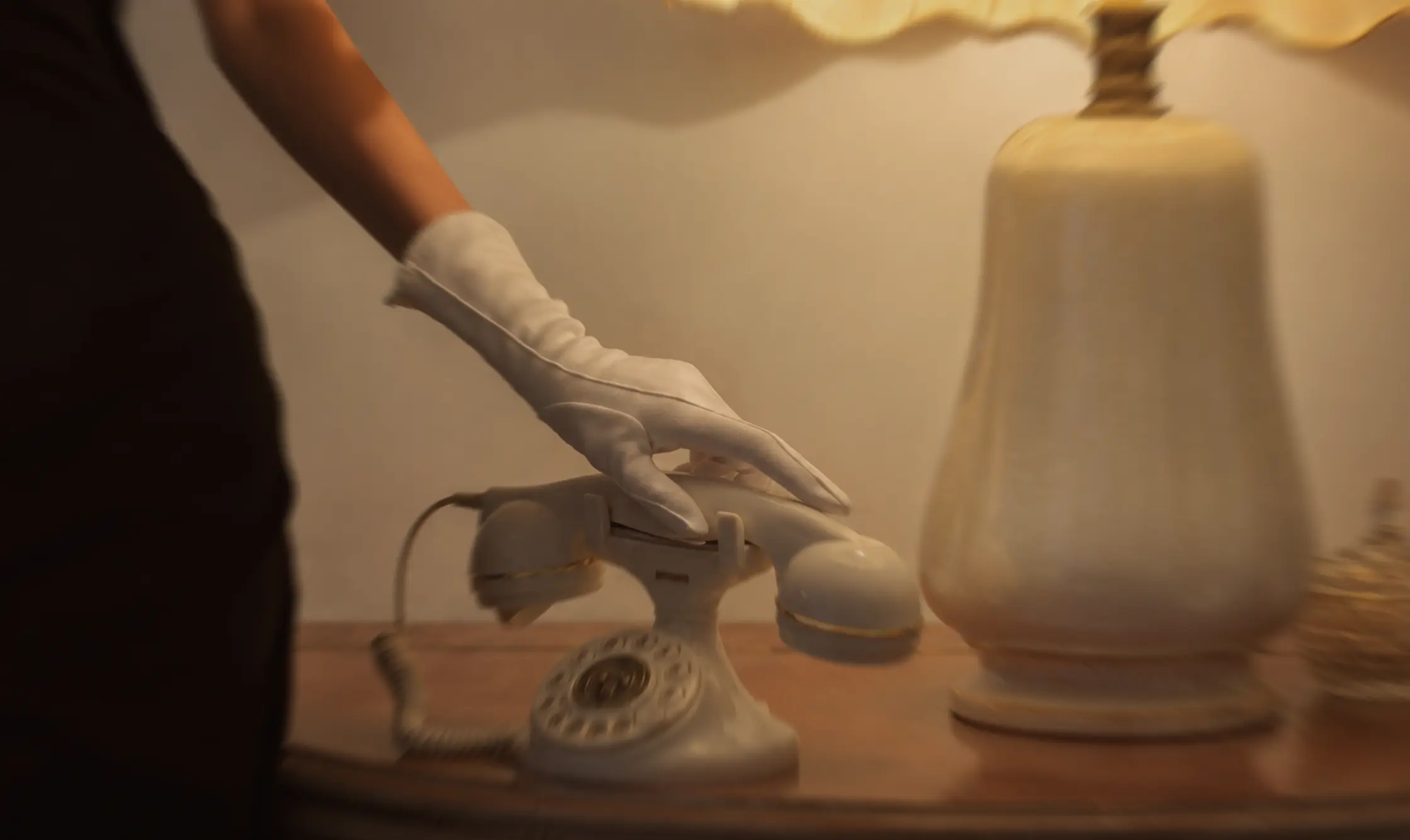 Hand in white glove reaching for a vintage rotary phone on a wooden table beside a lamp.